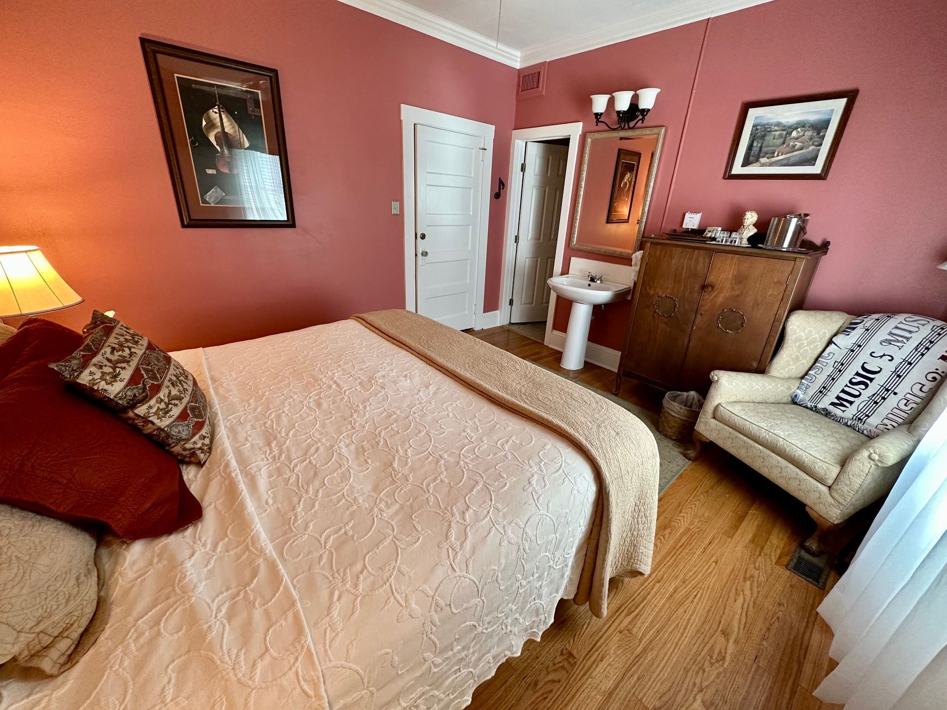 Bedroom with pink walls, a bed, armchair, chest of drawers, and artwork. A door leads to a bathroom with a pedestal sink.