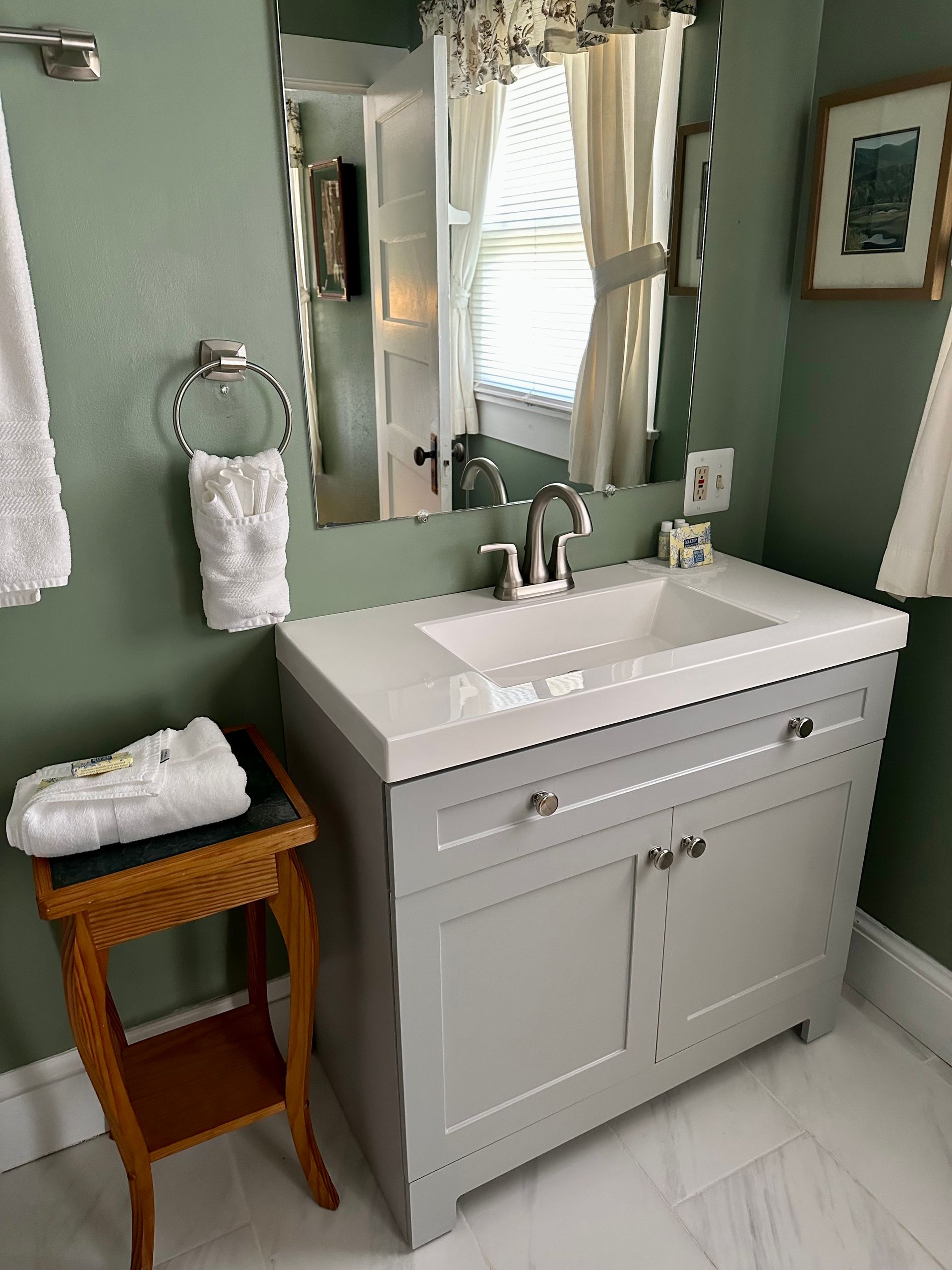 Bathroom with a gray vanity, white countertop, and sage green walls. A mirror reflects a window with curtains and a framed picture.
