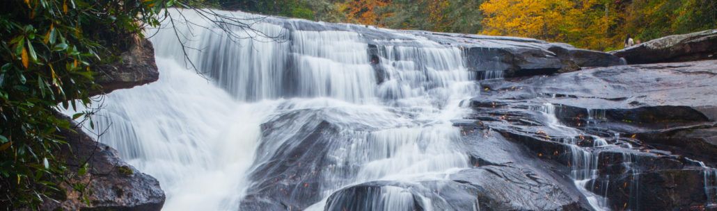 A waterfall cascades over layered rocks, surrounded by green and yellow foliage, suggesting an autumn scene.