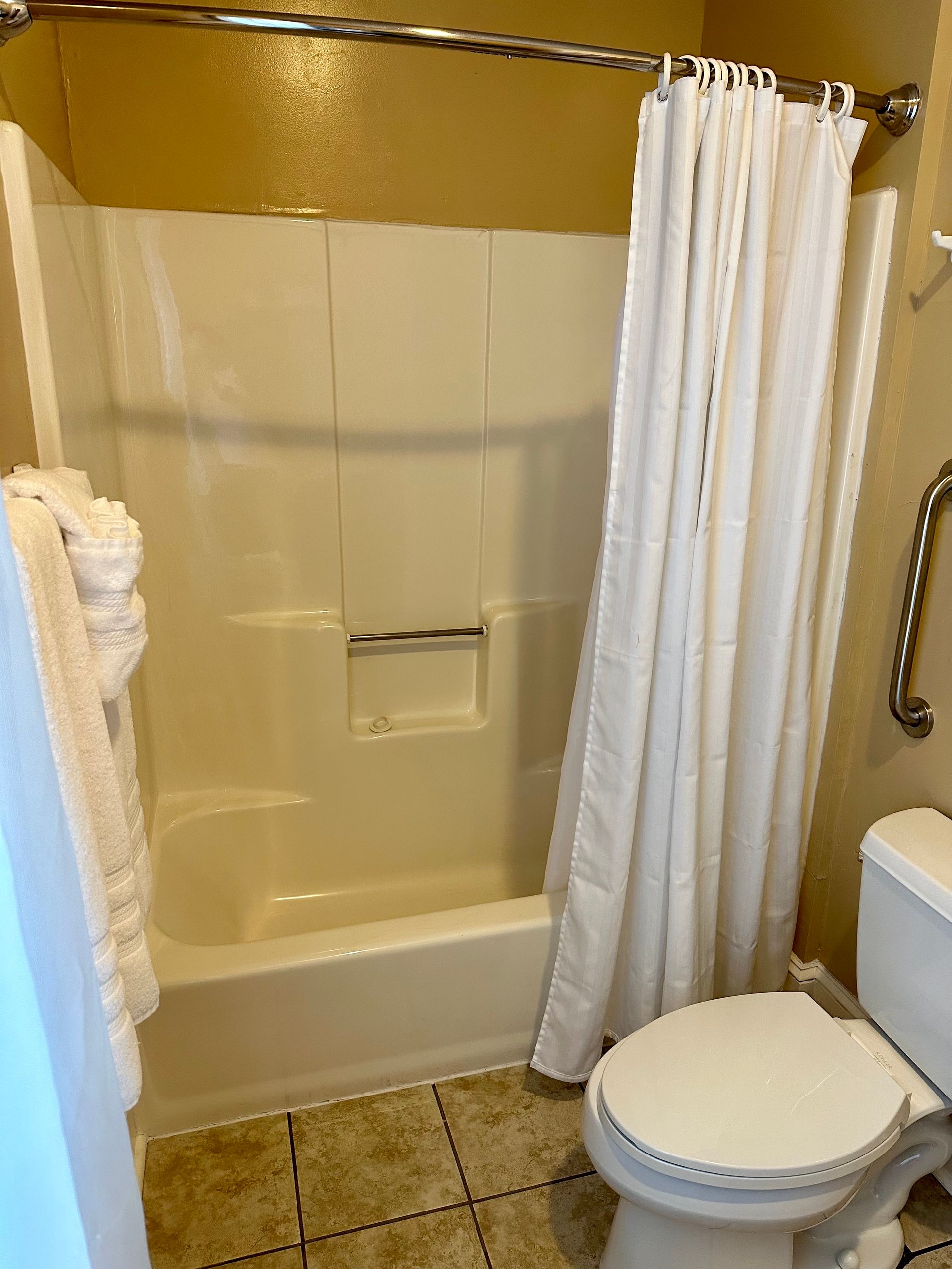 A small bathroom with a white shower, toilet, and a shower curtain. There is a grab bar next to the toilet.