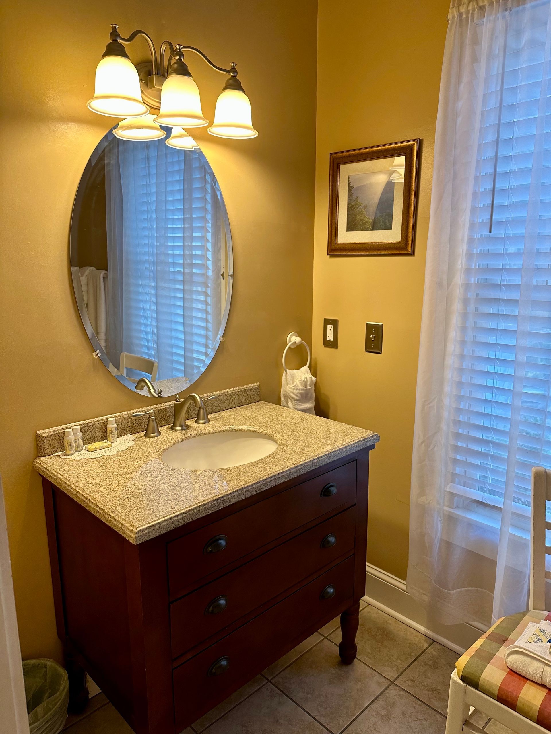 Bathroom with a red vanity, oval mirror, and three-bulb light fixture. Includes a window with blinds and a small framed picture.