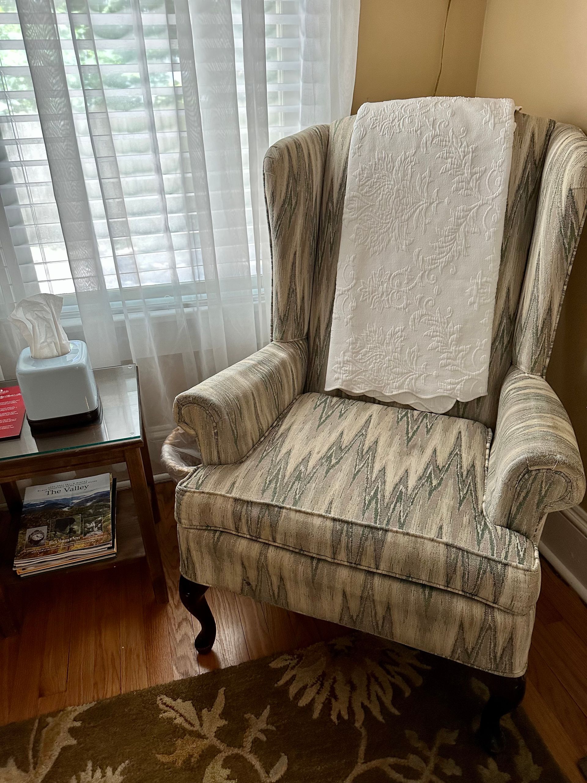 Wingback chair with patterned upholstery, a white throw, and wooden legs; beside a small table and window with blinds.
