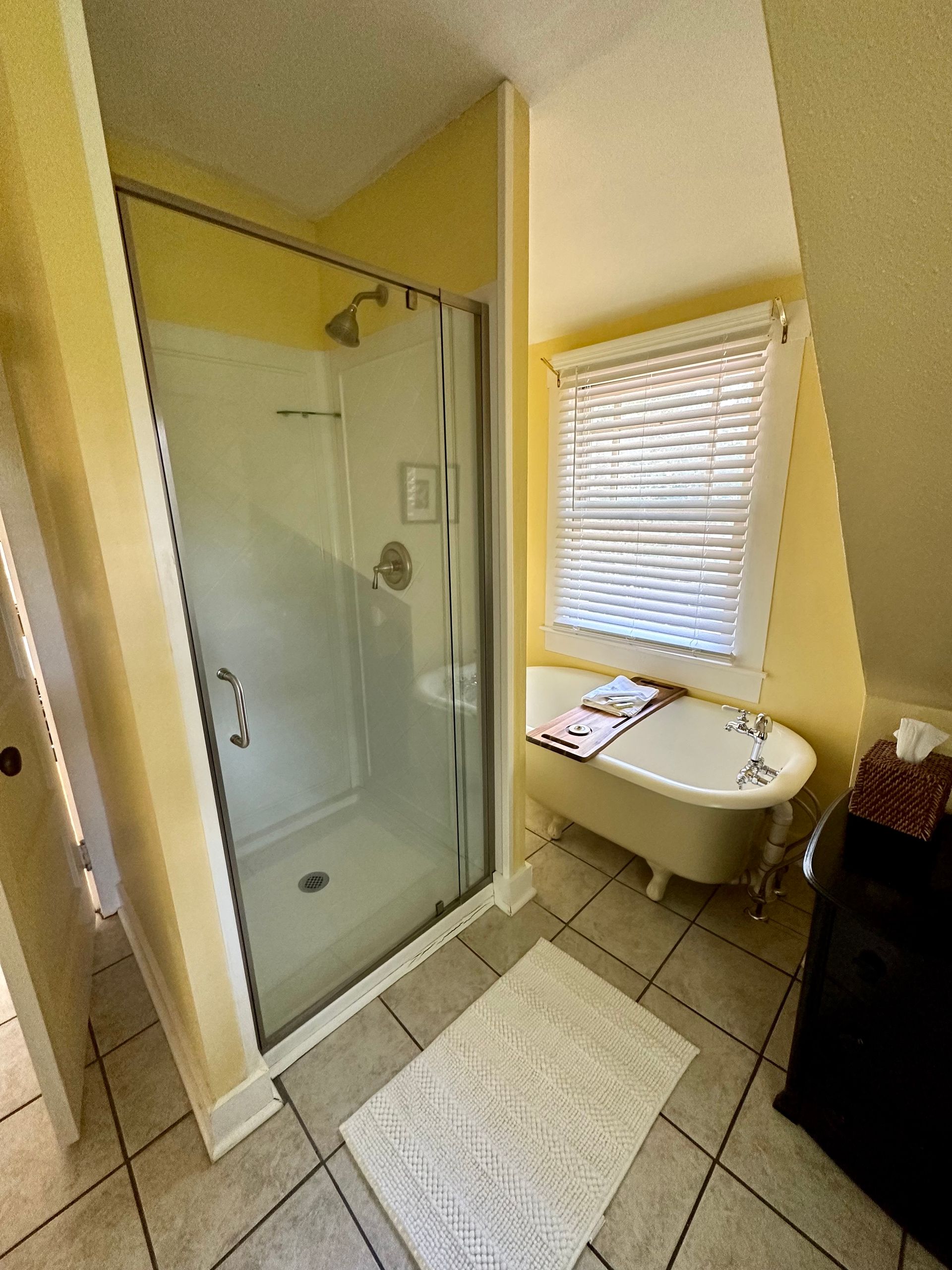 Bathroom with a glass shower, clawfoot tub, and window with blinds. Pale yellow walls and tiled floor.