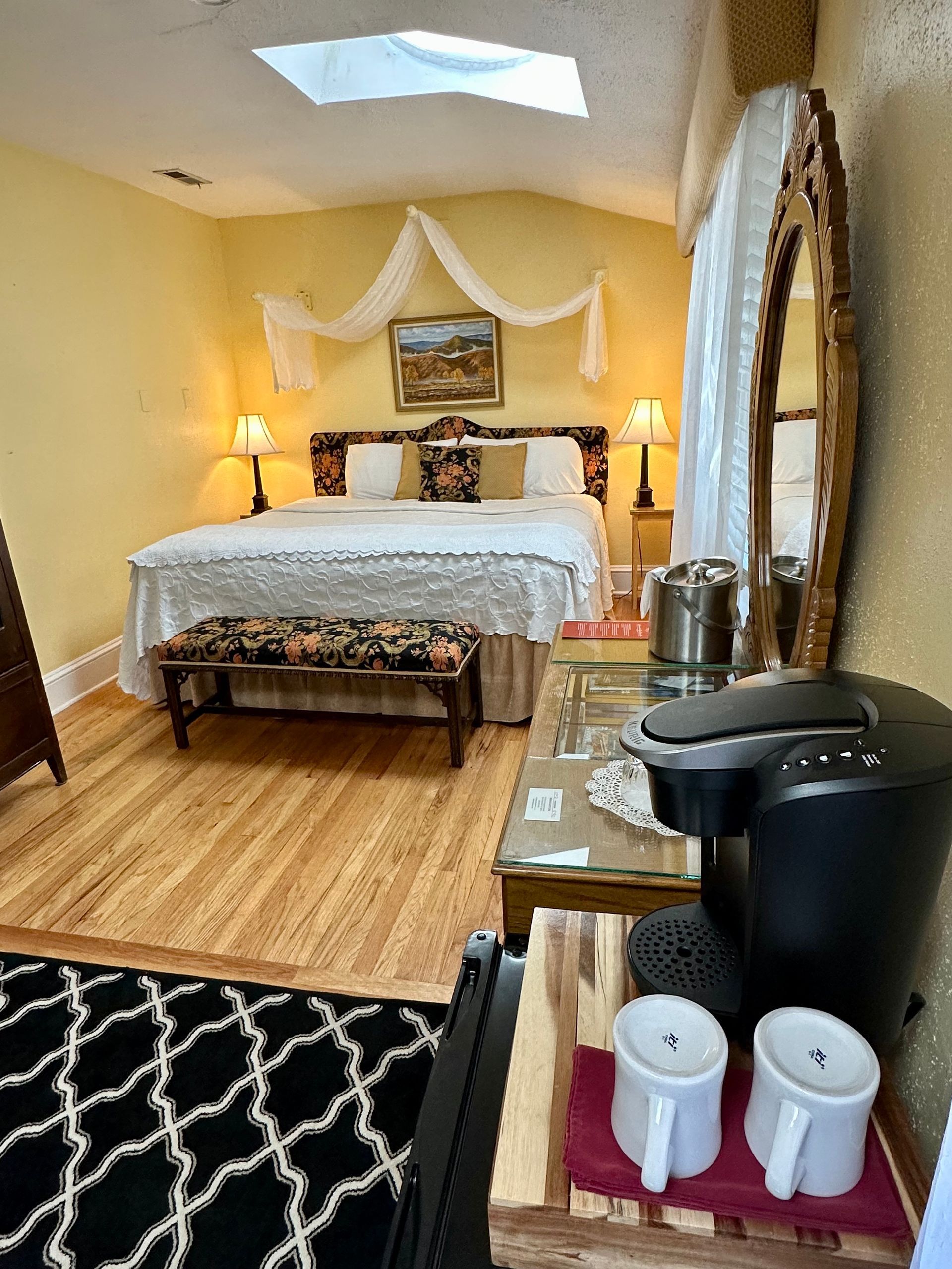 Cozy bedroom with bed, mirror, coffee maker, and patterned rug. Soft yellow walls, wooden floors, and natural light from a skylight.