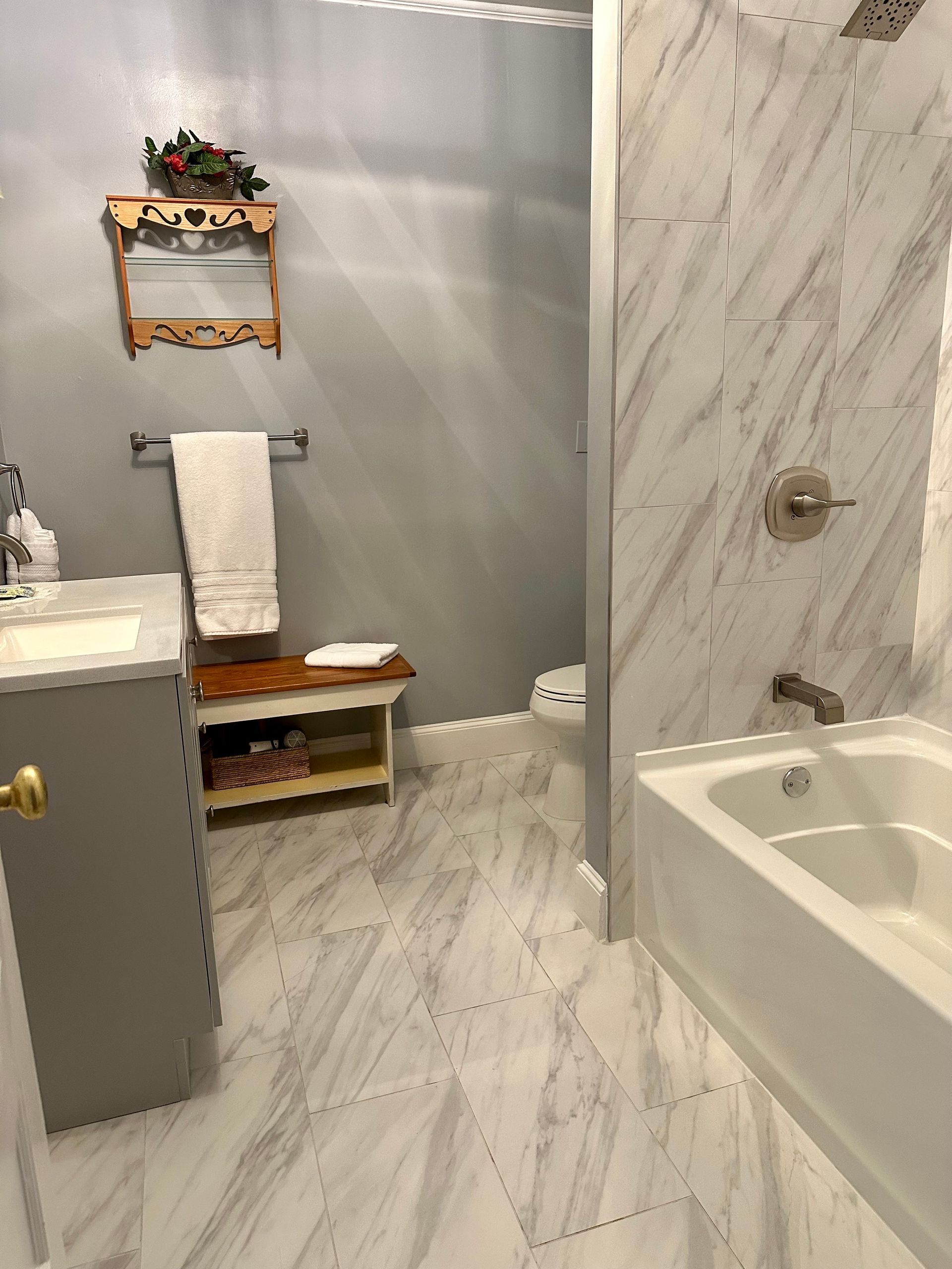 A bathroom with a marble-tiled floor and shower. Gray vanity and walls, with a wooden bench and shelf.