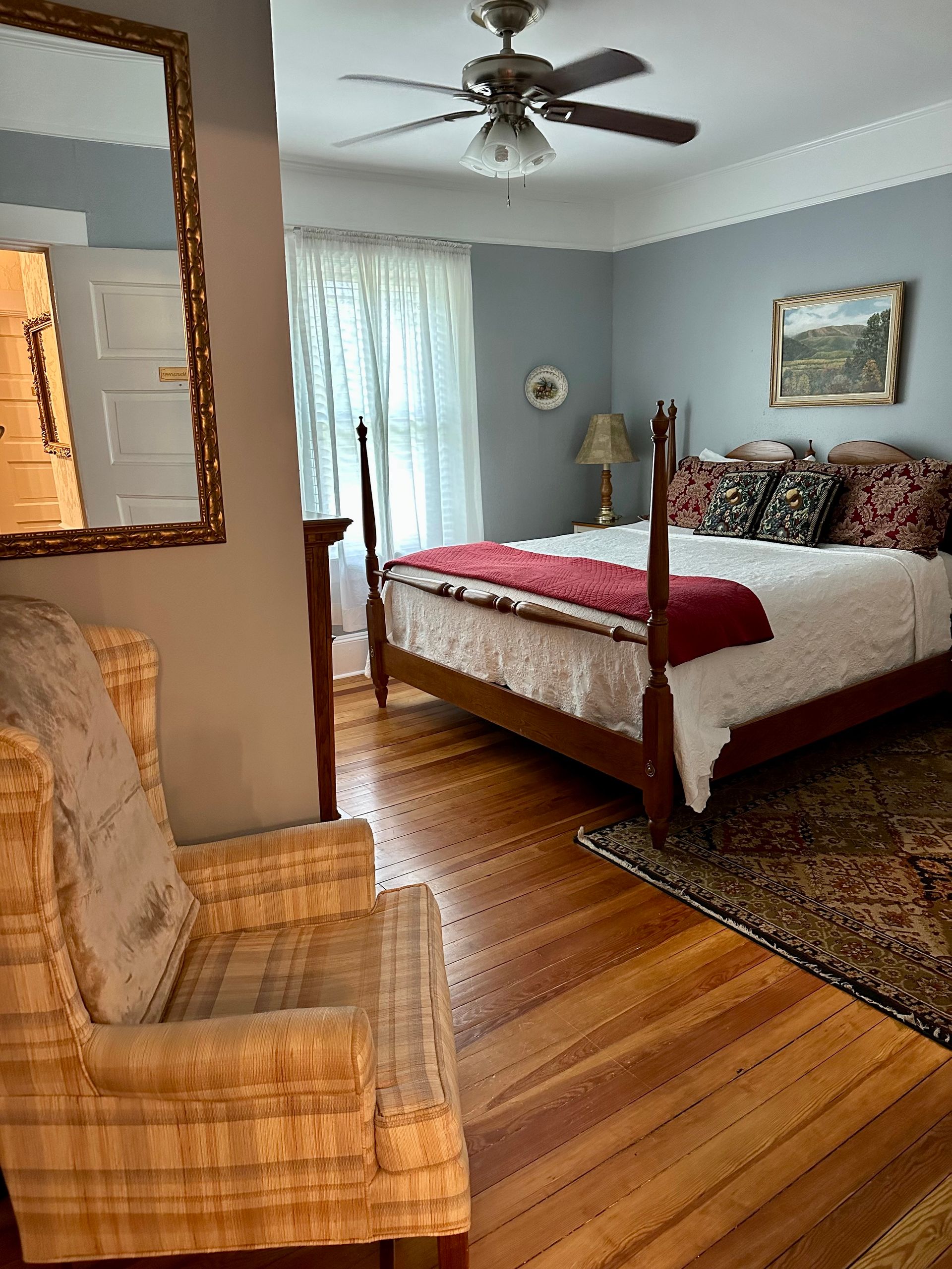 Bedroom with hardwood floors, a four-poster bed, and an armchair. The walls are pale blue, and the room has a vintage feel.