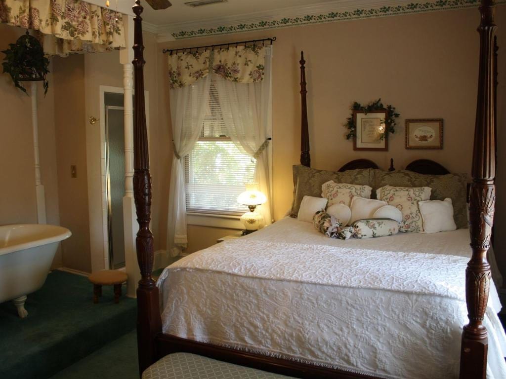 Bedroom with a four-poster bed, bathtub, and window. The room has floral decorations and soft lighting, giving a vintage feel.