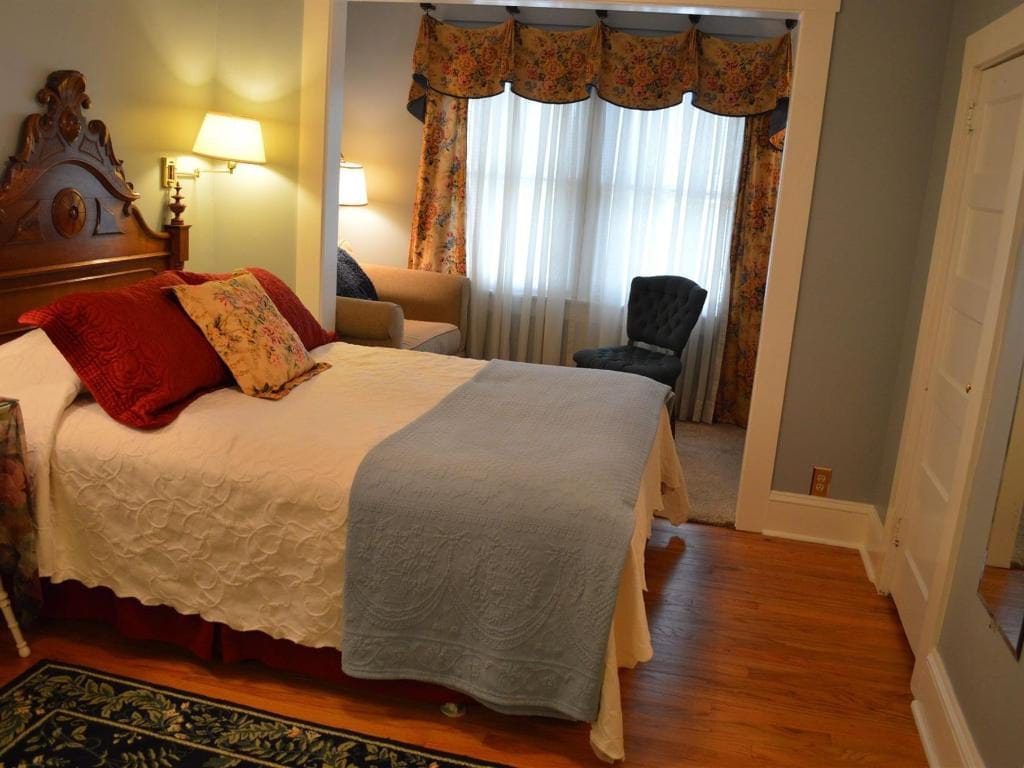 A cozy bedroom with a double bed, reading nook, and wooden floor. Features include a dark wooden headboard, red and patterned pillows, and a blue blanket.