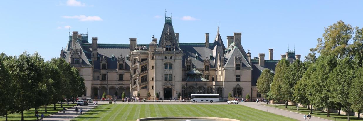 Biltmore Estate, a large stone mansion, with green lawns and trees, blue sky. A tour bus and people are visible in front of the building.