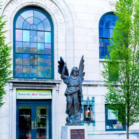 Bronze angel statue in front of Asheville Art Museum's entrance, a white building with arched stained glass windows and green trees.