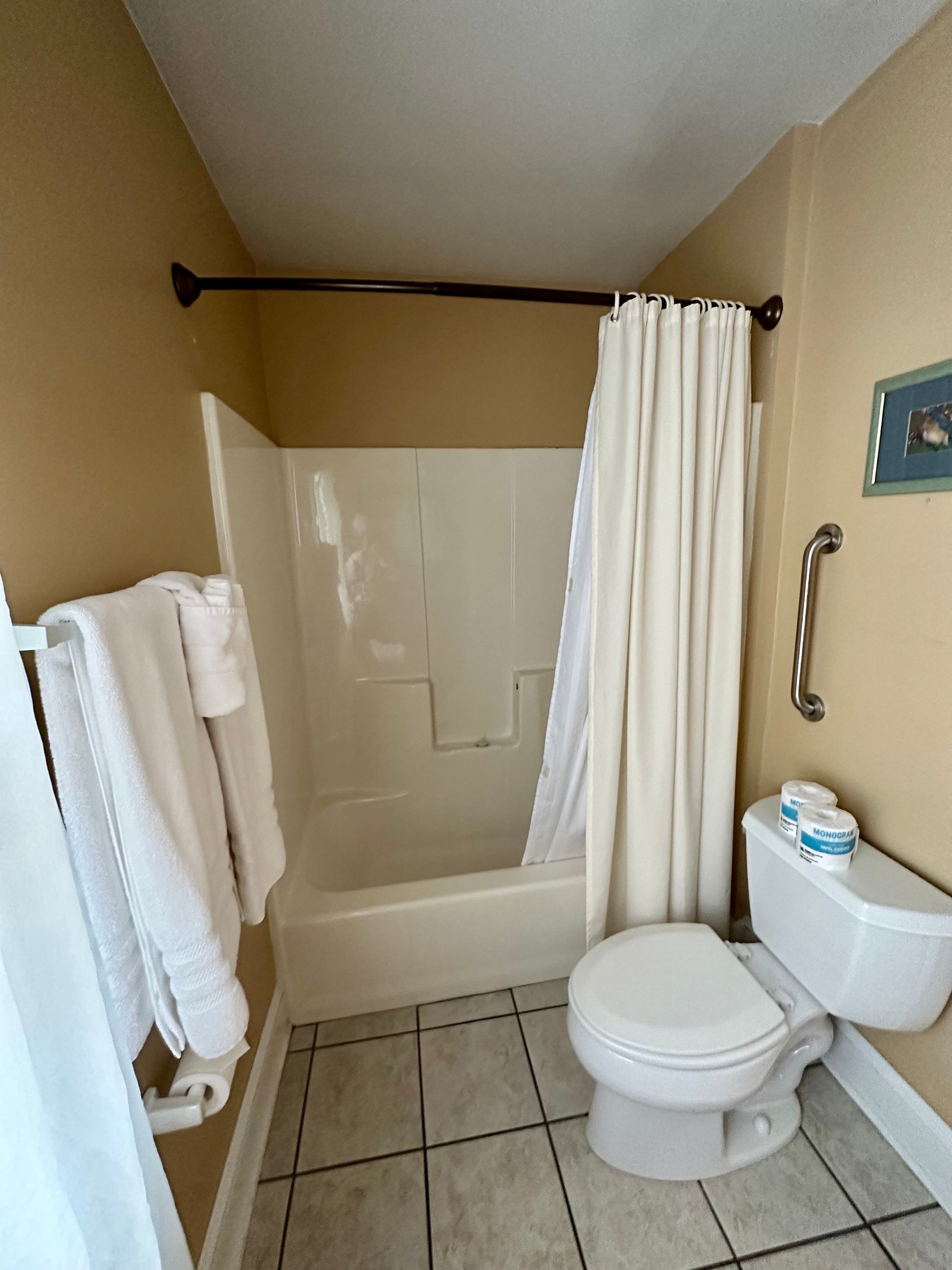 Bathroom with beige walls, a white shower/tub, a closed white curtain, a toilet, and a towel rack with white towels.