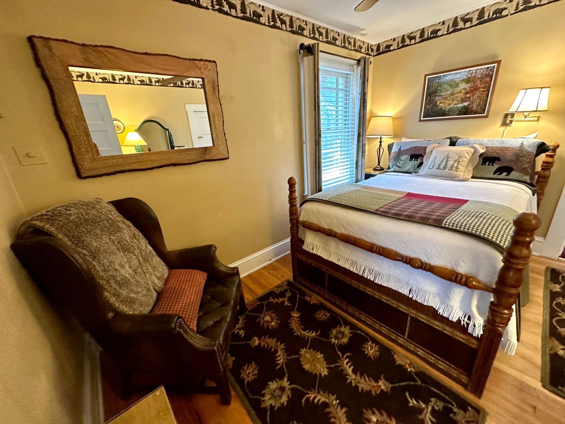 A bedroom with a wooden bed, armchair, mirror, and rug. The walls are beige with a decorative border.