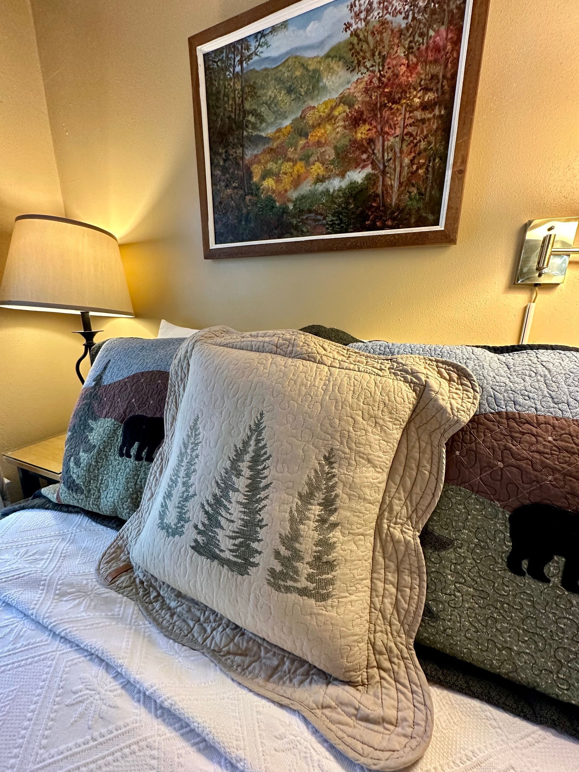 Bed with decorative pillows featuring trees and bears, under a framed landscape painting. Soft lighting, warm colors.