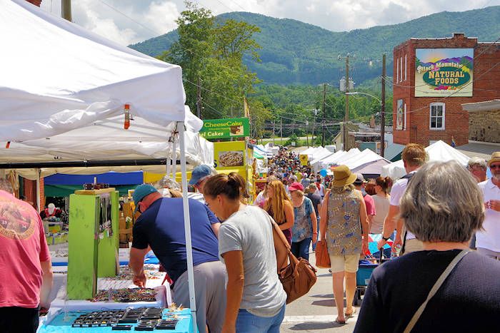 Outdoor market with crowds of people, white tents, and a backdrop of green mountains and a brick building.