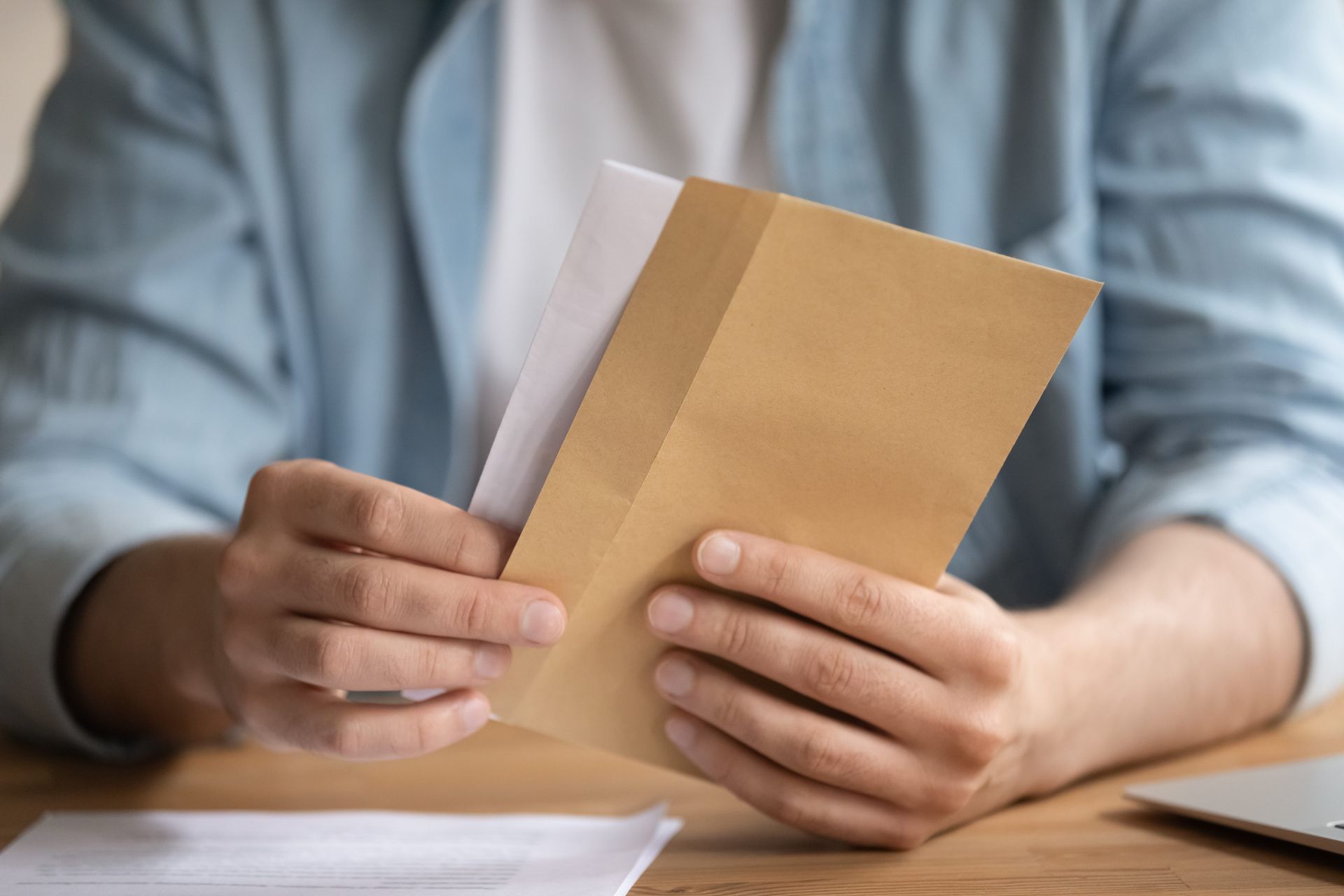 Hands holding and reading a tan envelope at a desk with papers and a laptop nearby