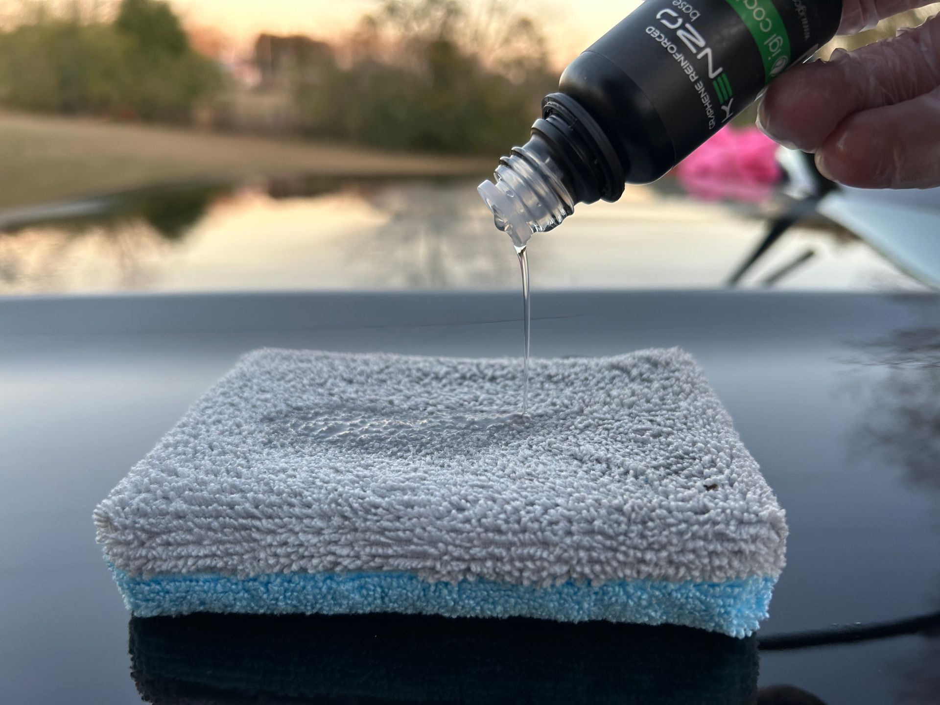 A bottle pouring liquid onto a sponge, likely for car detailing, on a black surface.