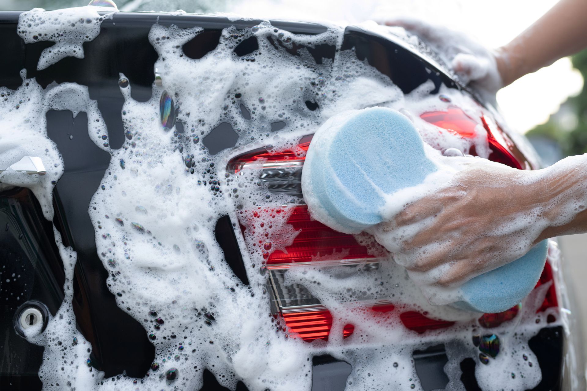 Person washing a black car with a blue sponge covered in soapy foam.