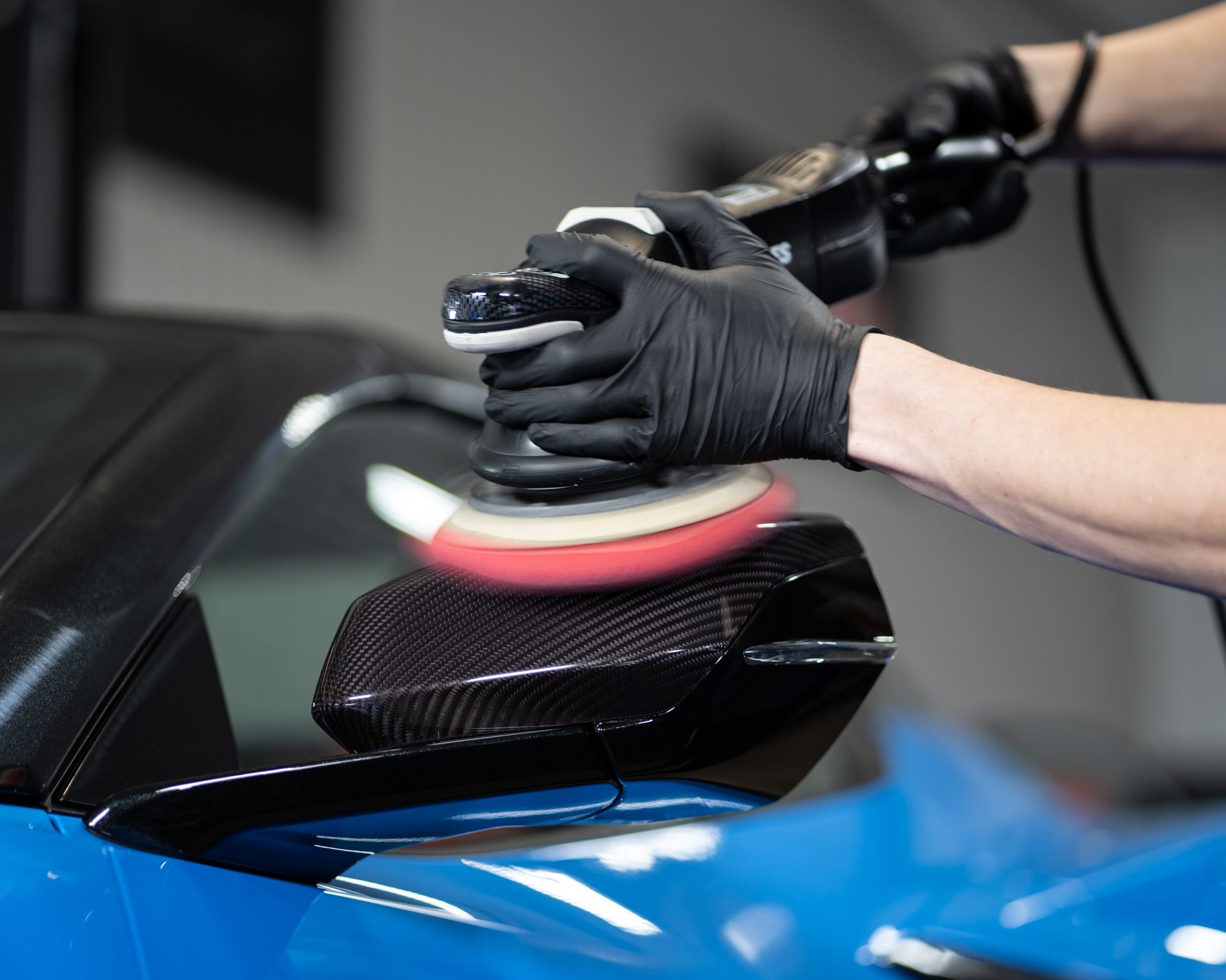 Person in black gloves polishing a blue car's black carbon fiber side mirror with an electric buffer.