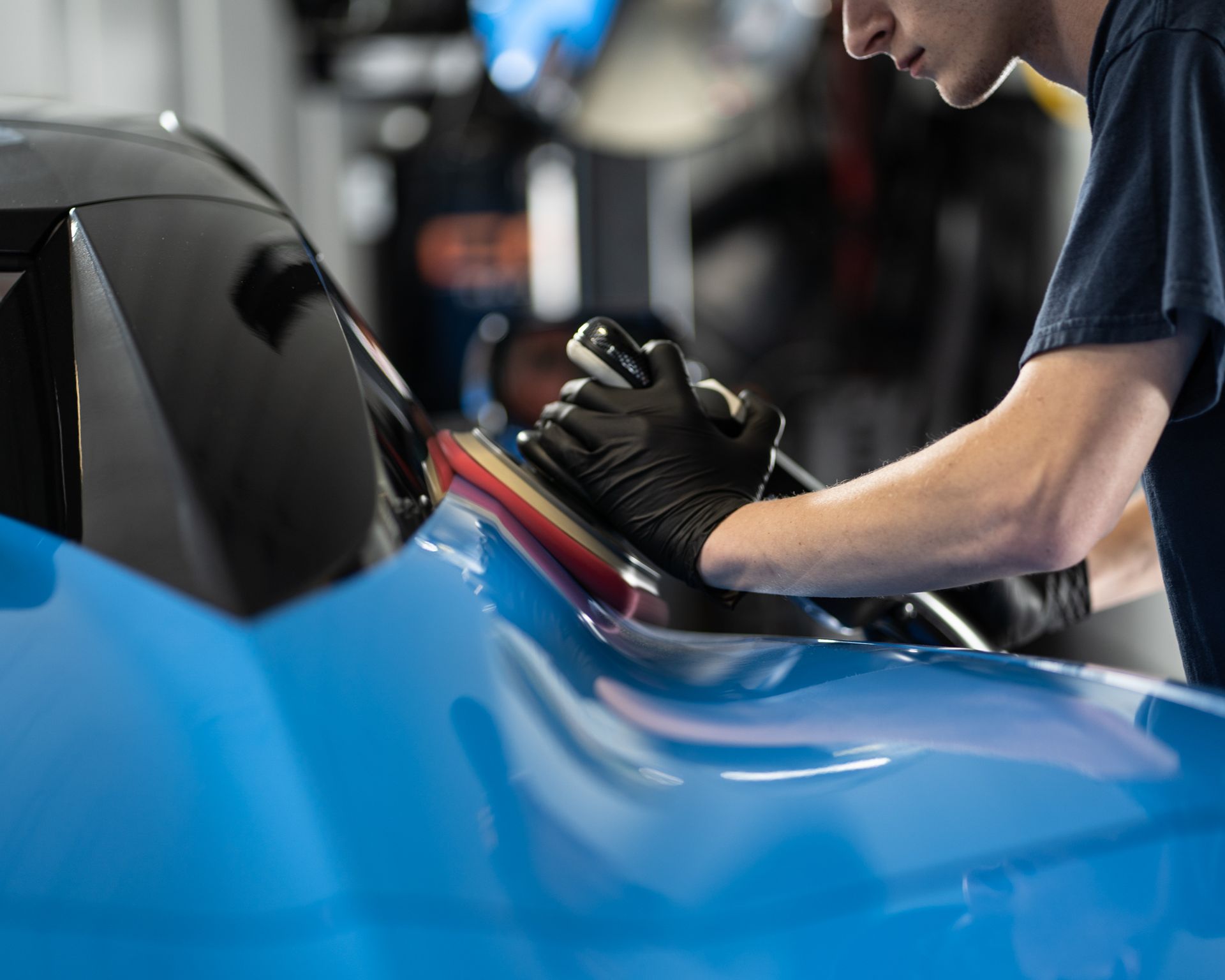 Person polishing a blue car with a machine and wearing black gloves.