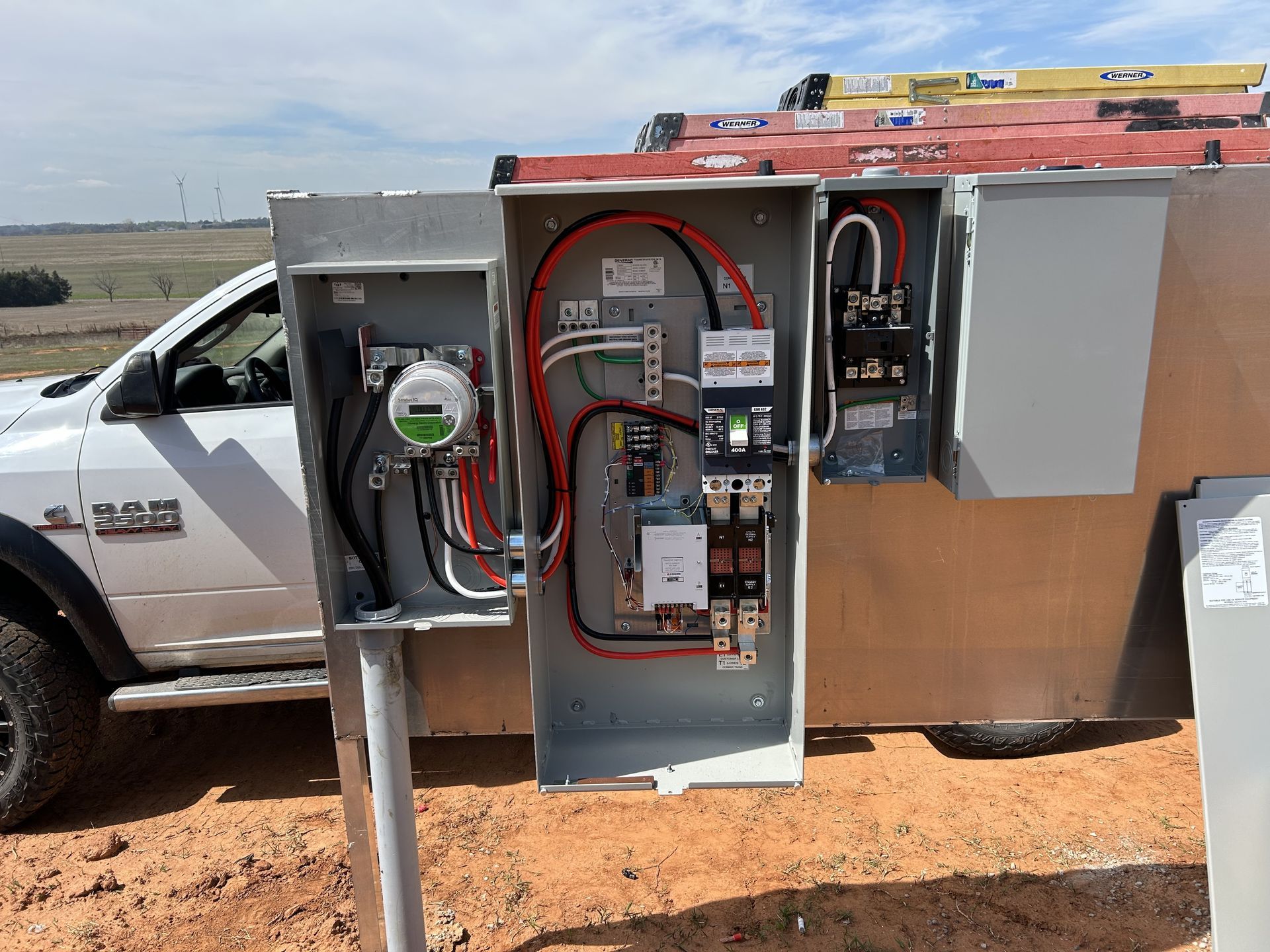 Electrical panel with exposed wires, meter, and breakers, mounted on a metal pole outdoors, next to a white truck.