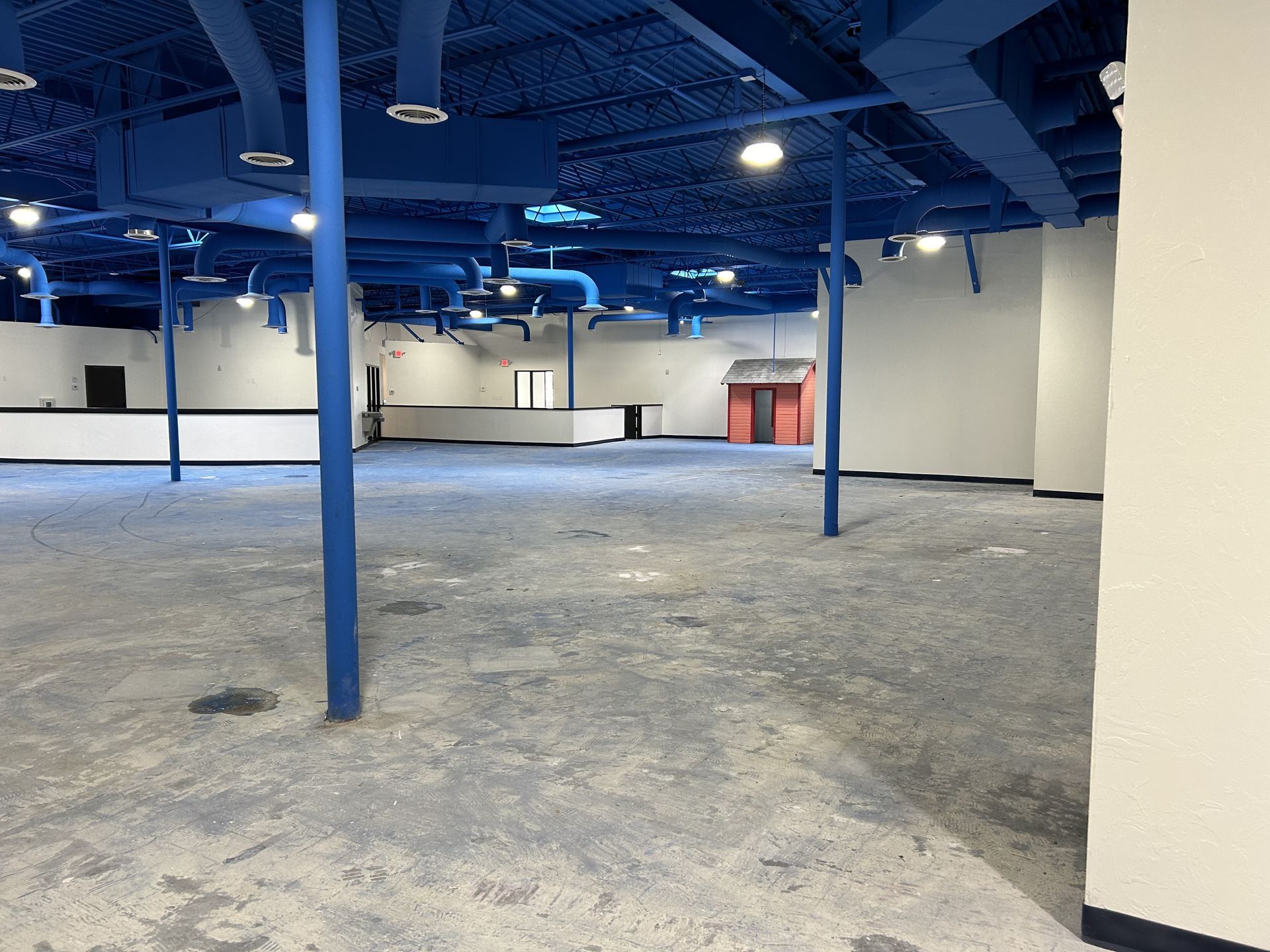 Empty room with blue ceiling and supports, gray floor, white walls, and a small red playhouse.