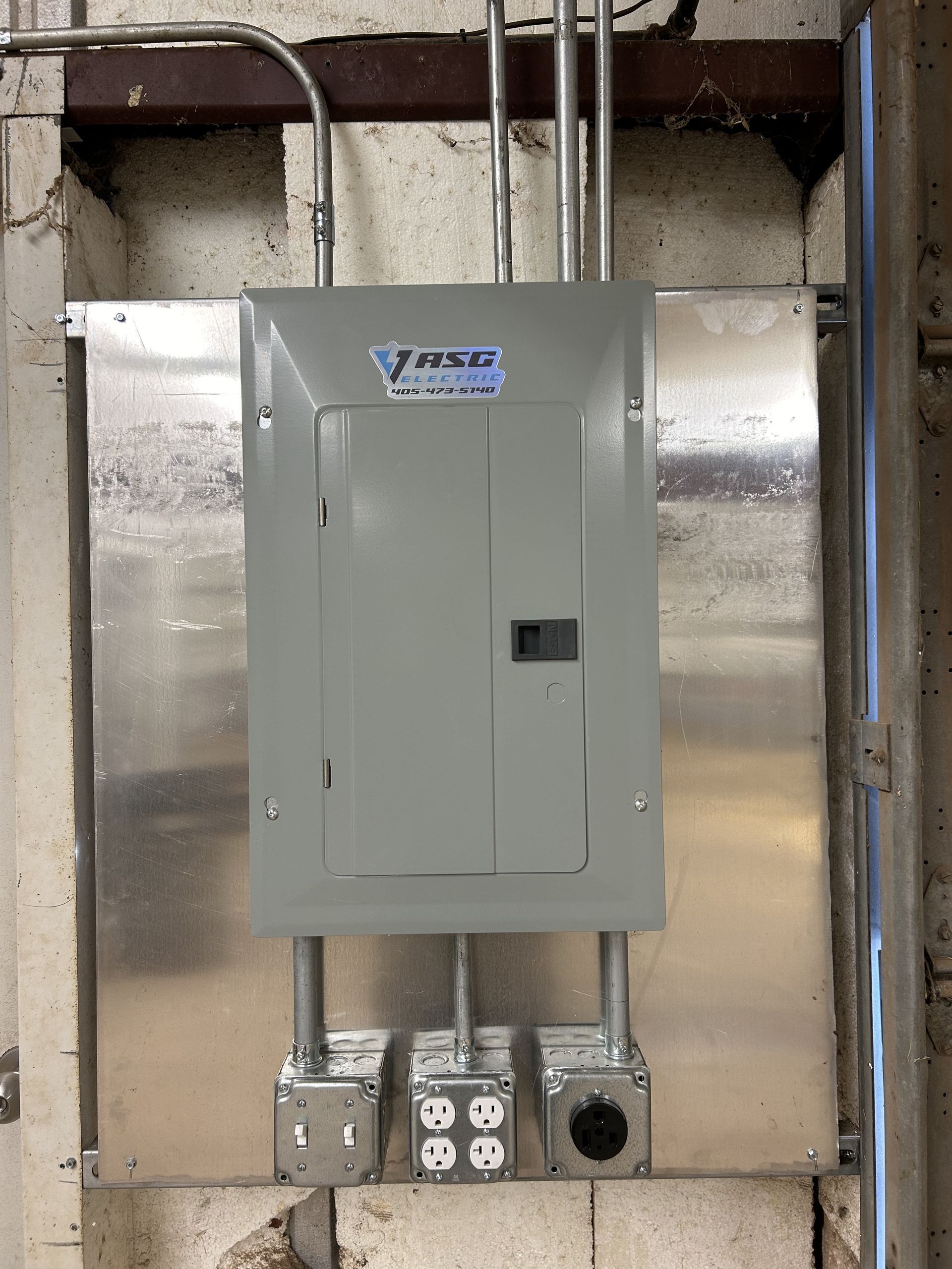 Electrical panel with conduits and outlets mounted on a metal plate. Gray panel, light-colored wall.
