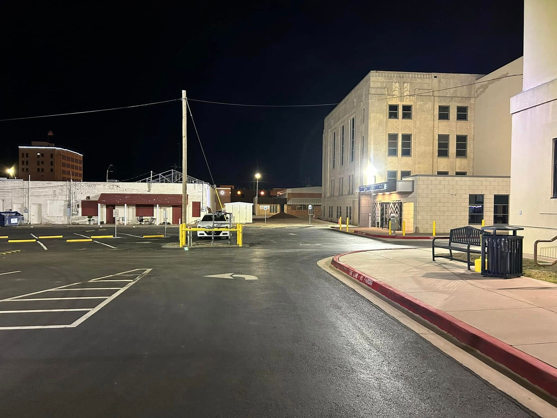 Nighttime view of a parking area, buildings, and streetlights. Paved road with painted parking spaces.
