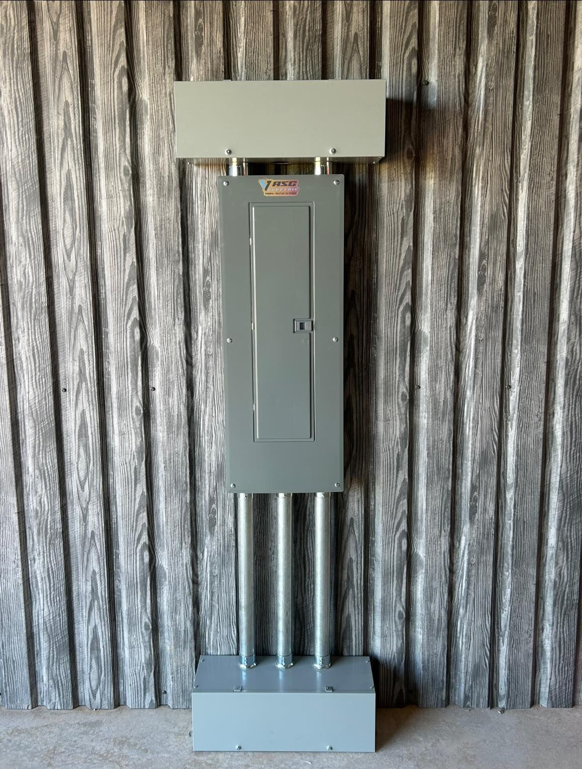 Gray electrical panel with conduit pipes, mounted on a base, against a wood-paneled wall.