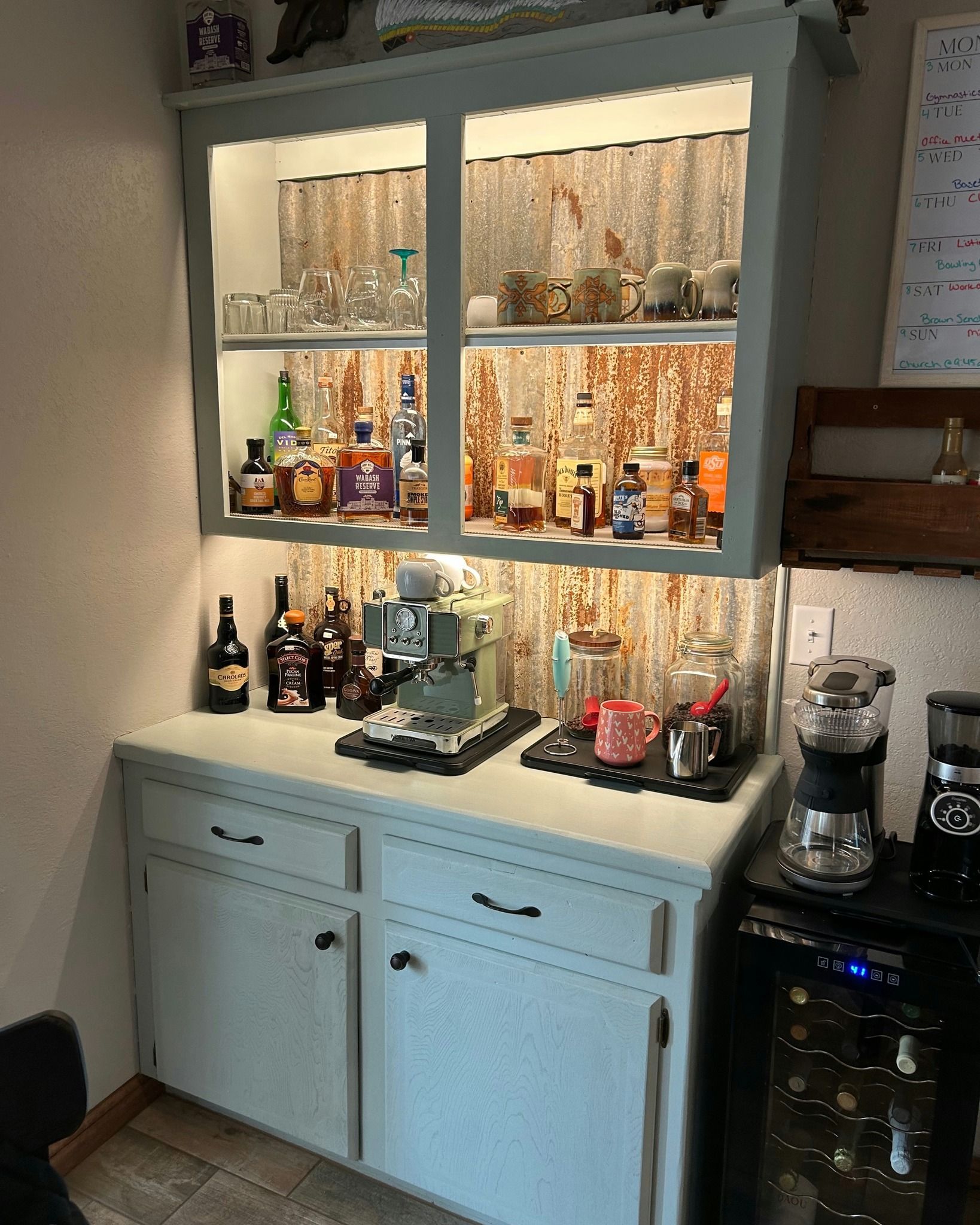 Light blue cabinet with liquor and coffee bar setup.  Top cabinet with glassware and bottles; lower cabinet, espresso .