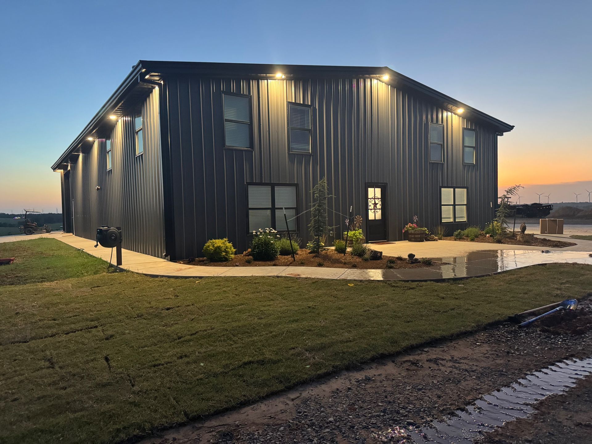 Black metal building with lit exterior lights at dusk. Green grass and stone patio in front.