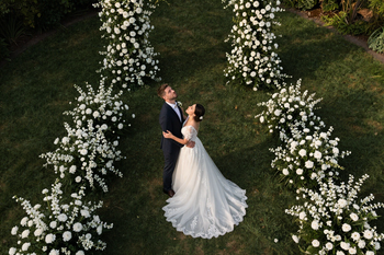 La novia y el novio, vestidos de blanco y negro, están de pie en un pasillo flanqueado de flores 