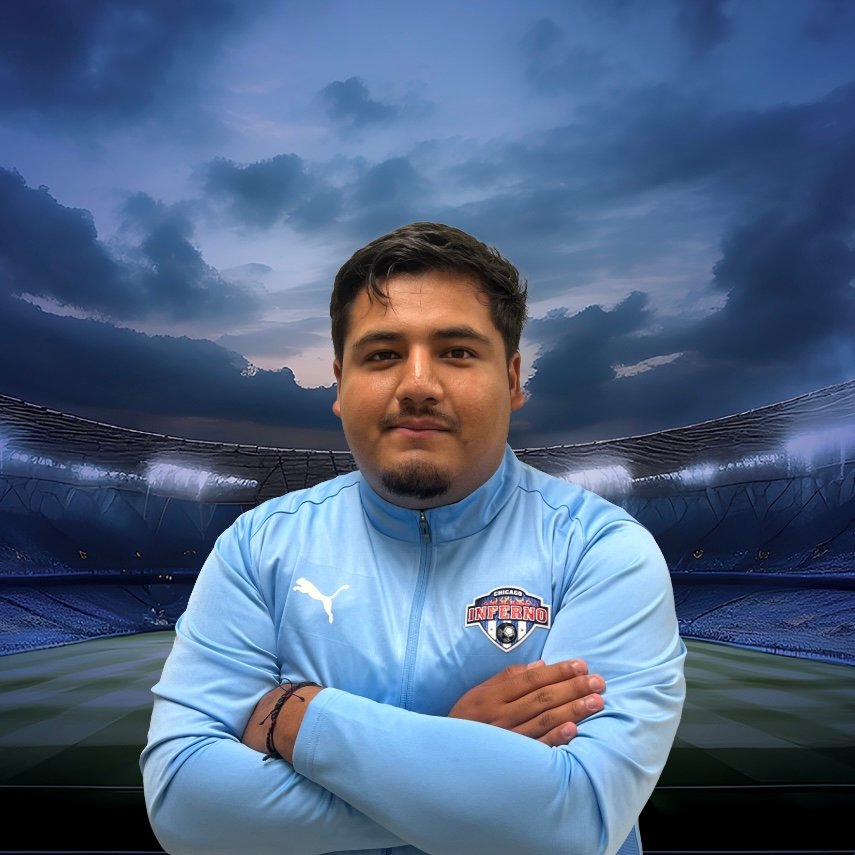 Coach Cristo Varela in light blue jacket, arms crossed, stands in front of a soccer stadium backdrop.