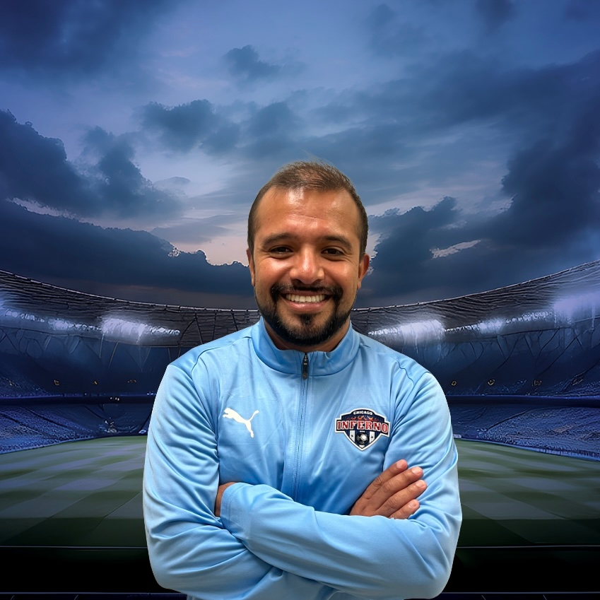Coach Carlos Alvarez in blue jacket with arms crossed, smiling, in front of stadium background.