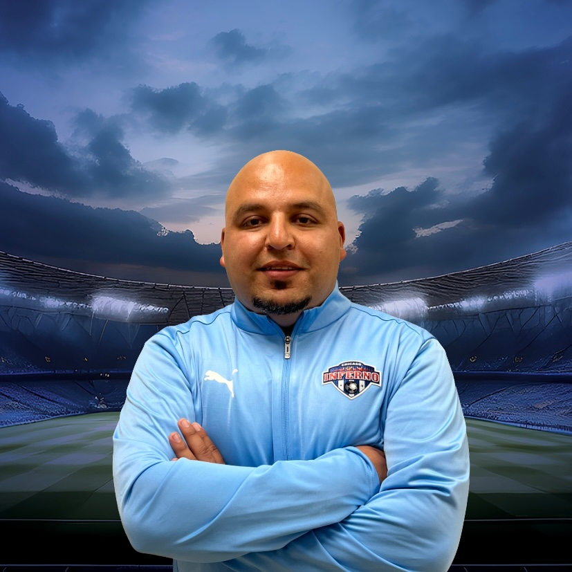 Coach Chuy Cruz in light blue jacket with arms crossed, standing in front of a stadium background.