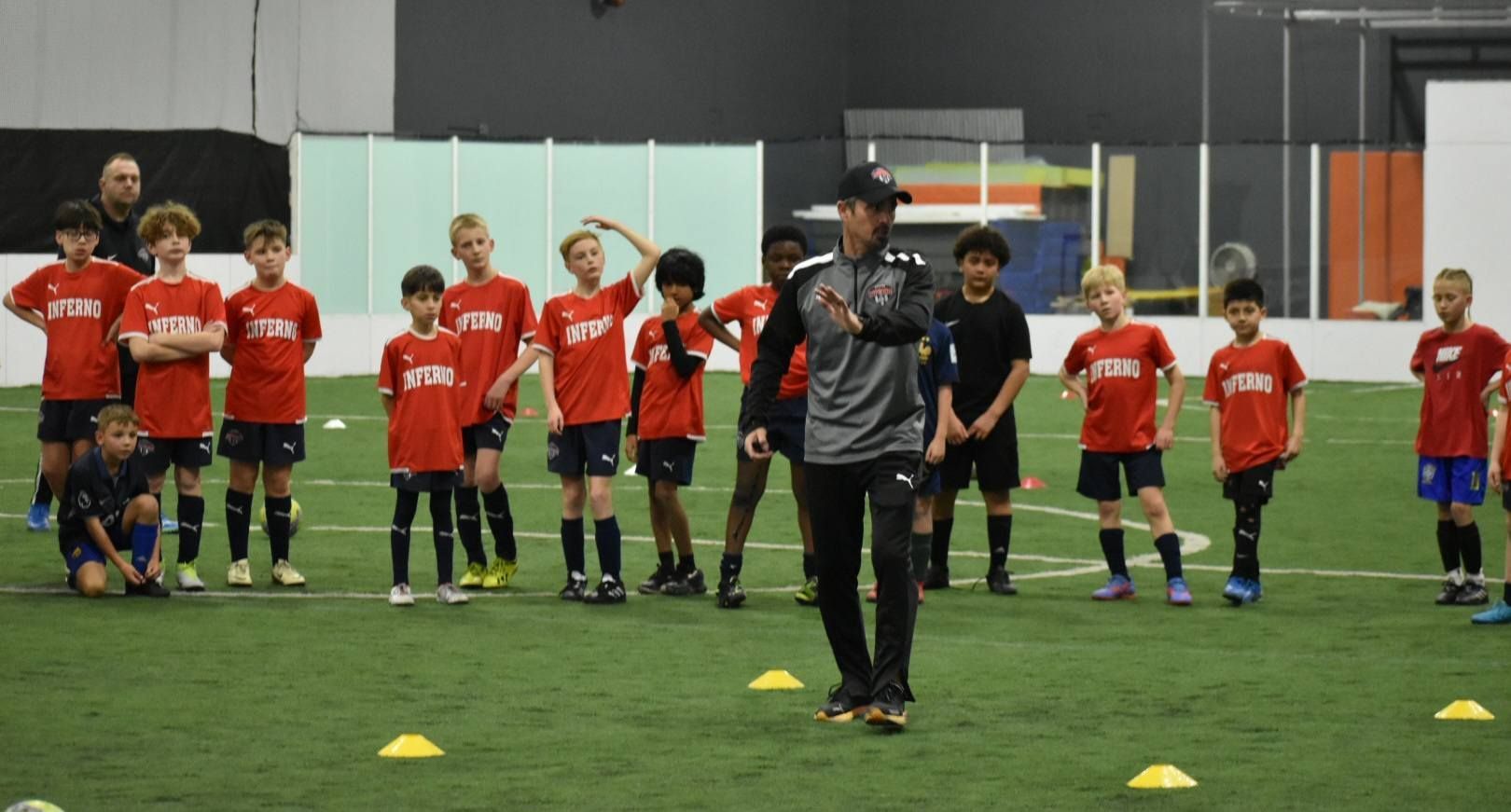 Coach instructing a youth soccer team on an indoor green turf field. Players wear red and black.