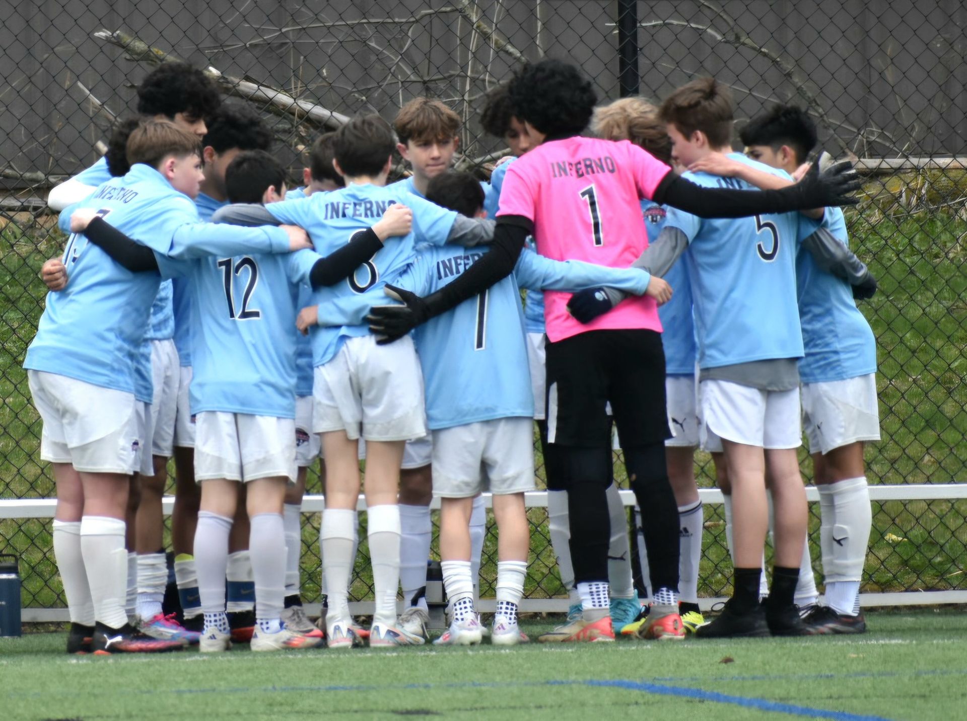 Soccer team in light blue jerseys huddled on a field, arms around each other.