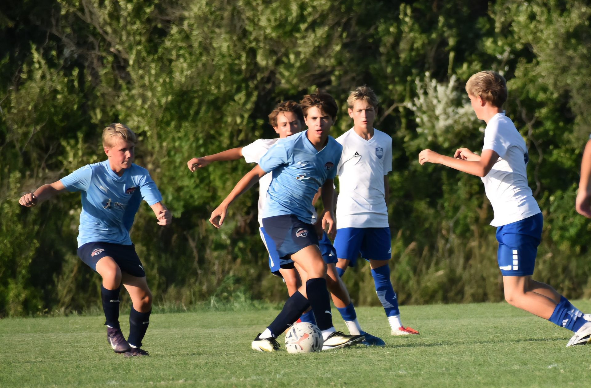 Soccer players in light blue uniforms on a green field; one dribbles the ball as others defend.