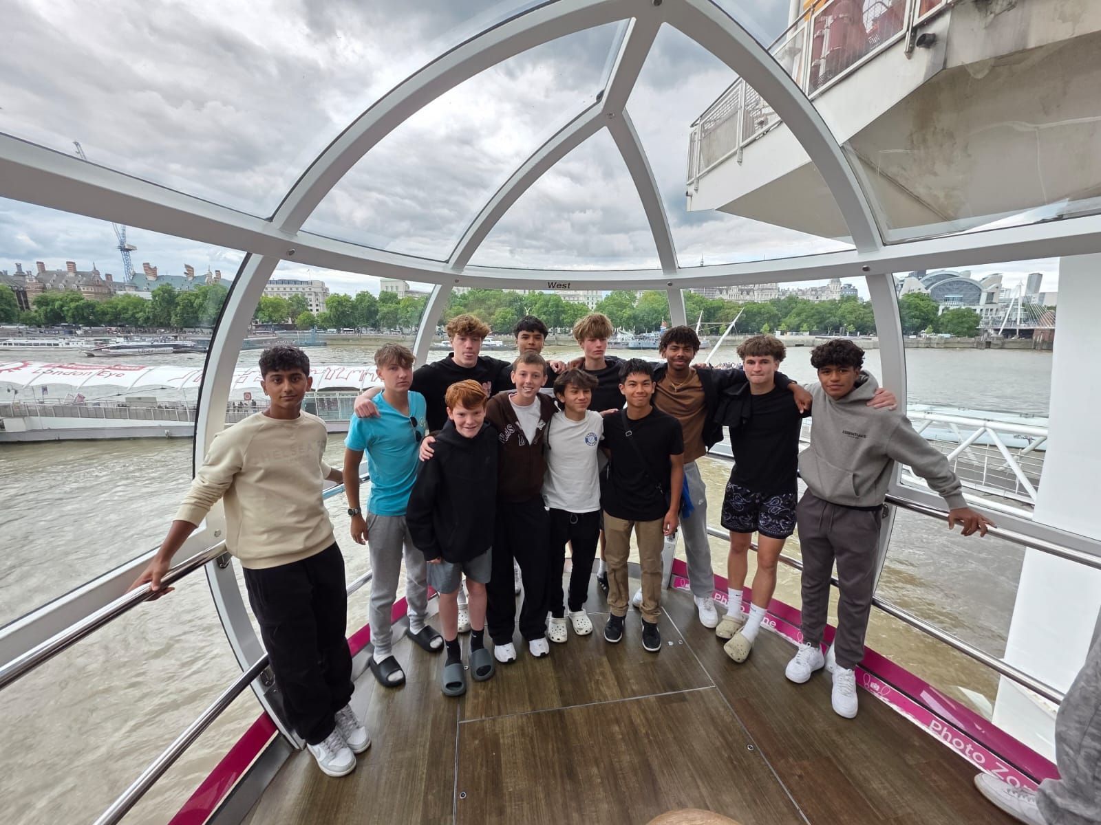 Group of teens inside a glass structure, overlooking a river. They are smiling and posing together.