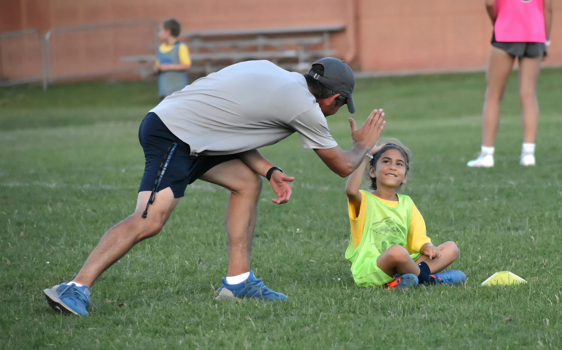 Man giving a high-five to a girl sitting on grass. They are on a soccer field, with others in the background.