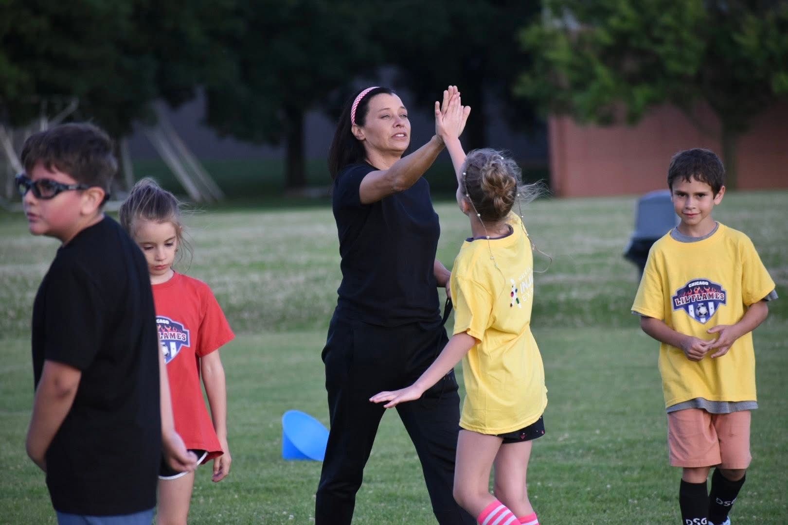Children outdoors playing a game, with  Coach Glory in black high fiving in the center.