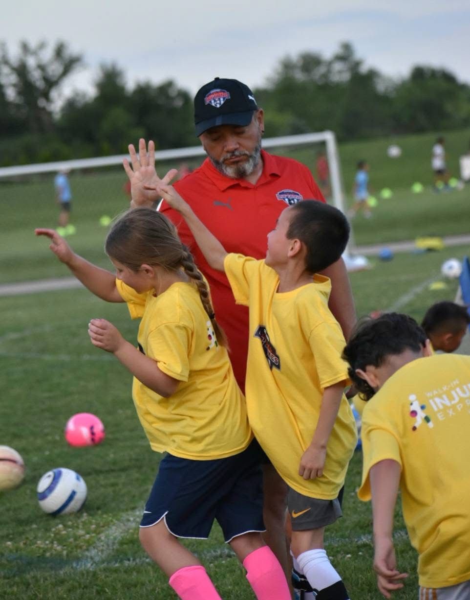 Soccer coach high-fives two young players in yellow shirts on a field, others nearby.