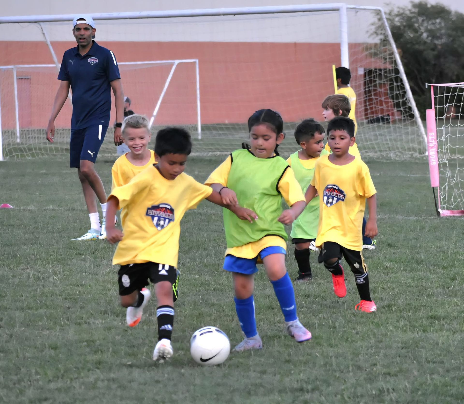 Youth soccer game: children in yellow shirts playing, coach watches, sunny outdoor field.