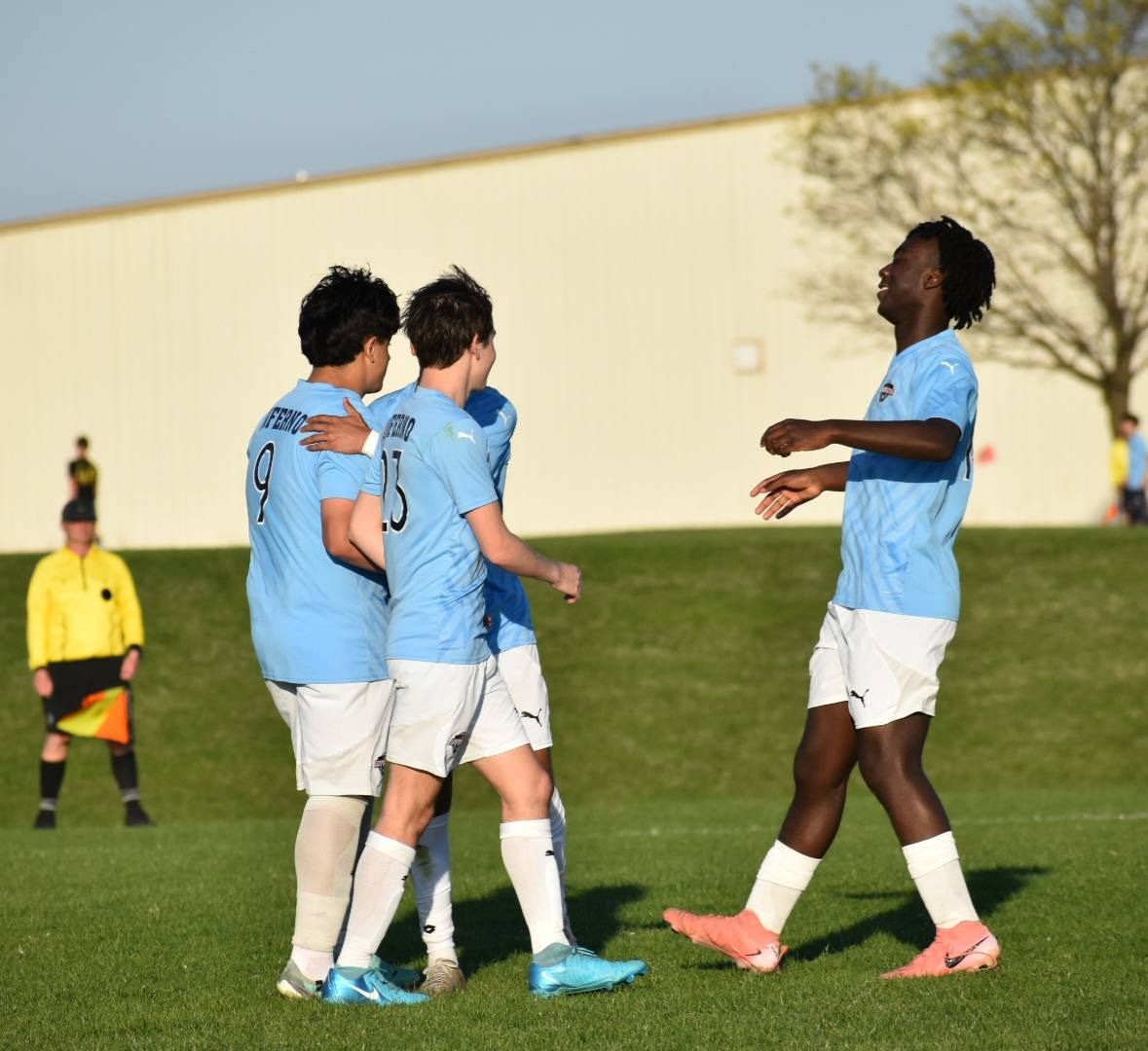 Soccer players in light blue jerseys celebrate on a green field. A referee watches in the distance.