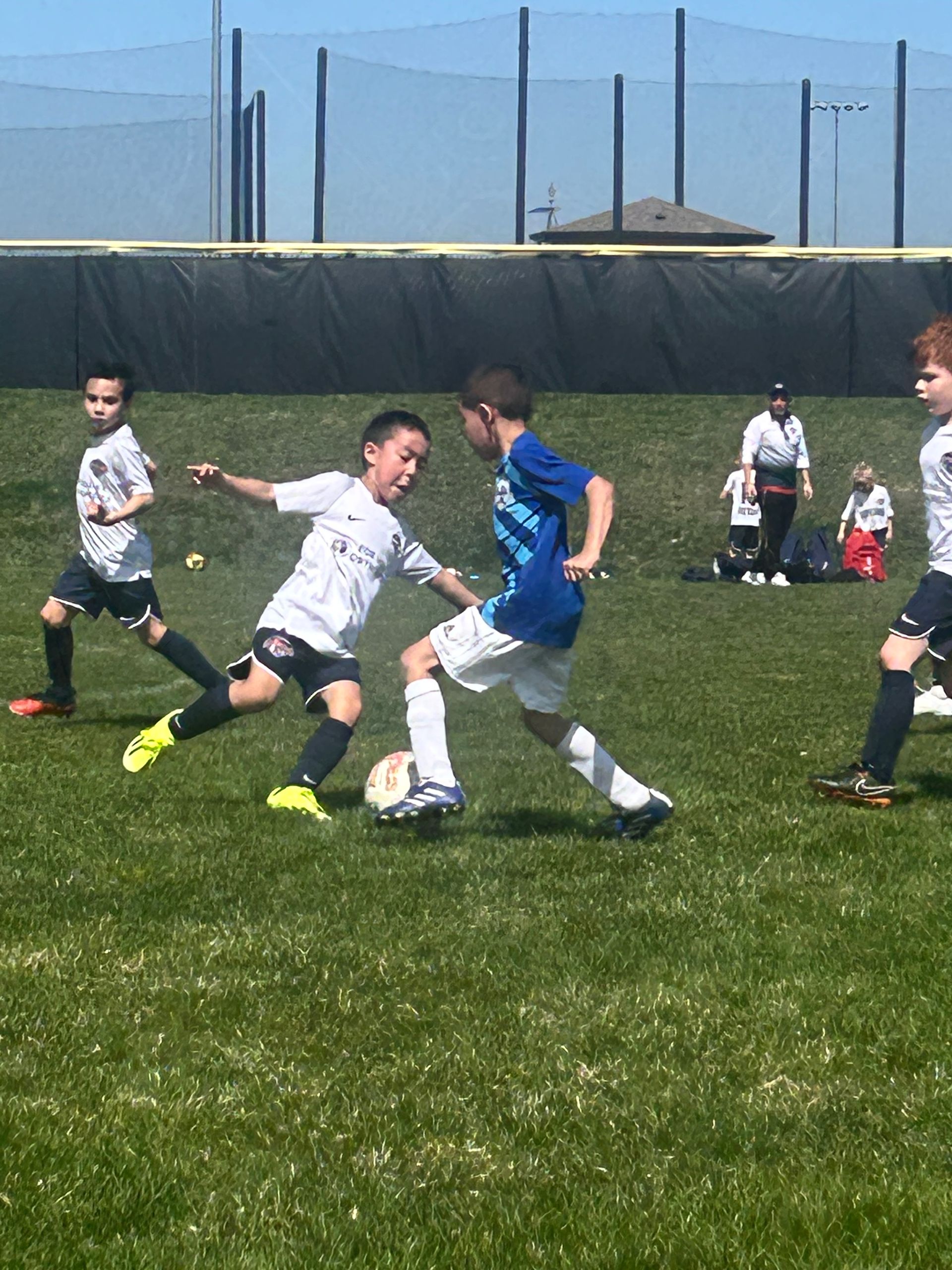 Two boys playing soccer on a grassy field; one in white, one in blue, kicking for the ball.
