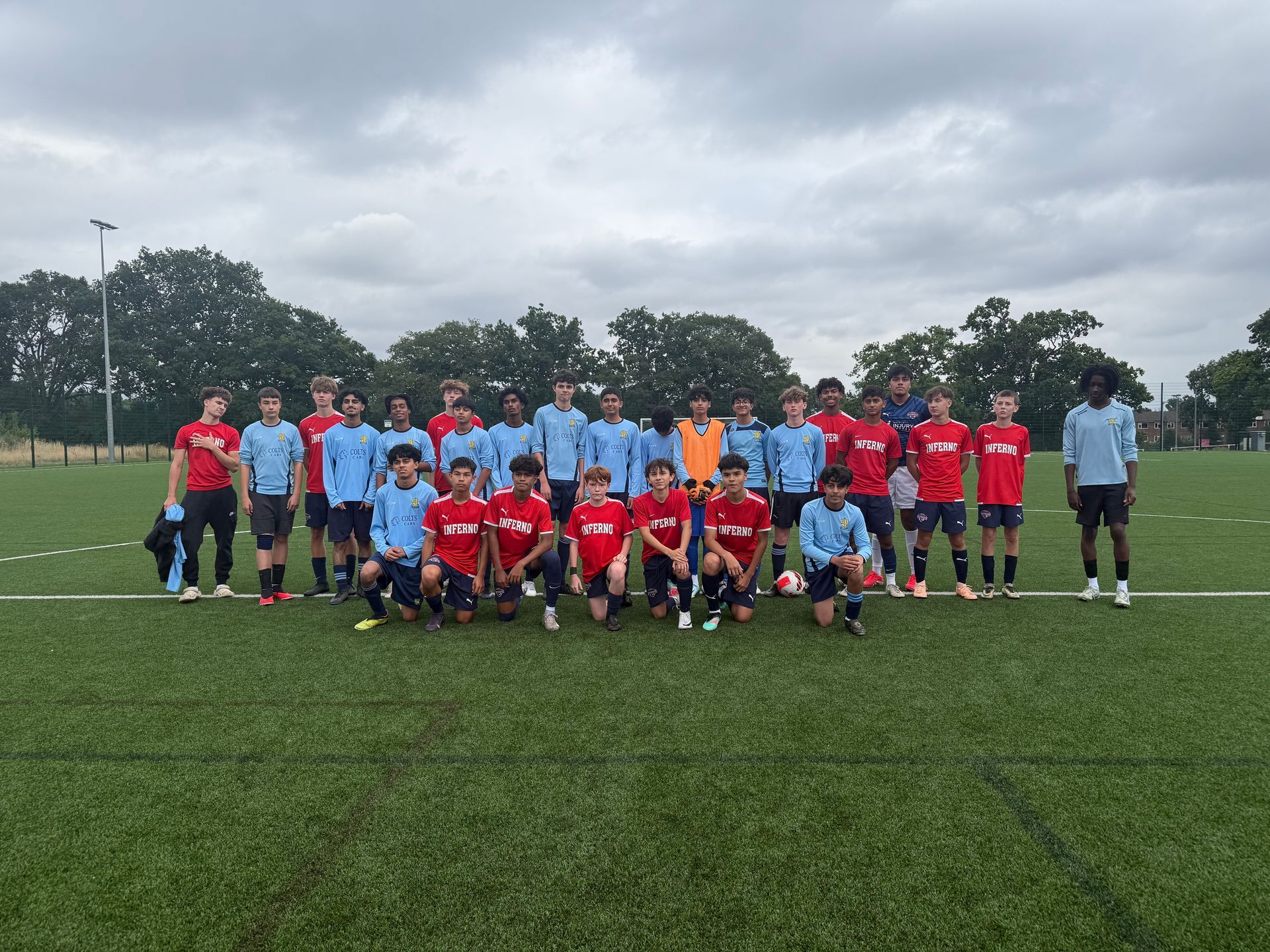 Soccer teams in red and blue uniforms pose together on a green field under a cloudy sky.