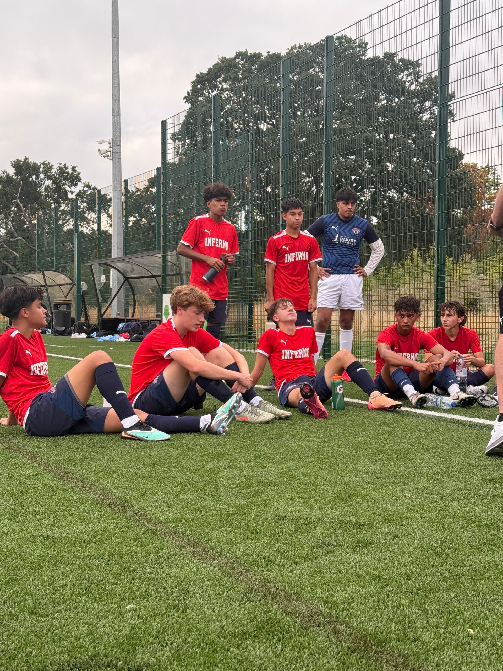 Soccer team in red shirts resting on a green field. Some sitting, others standing, near a fence.