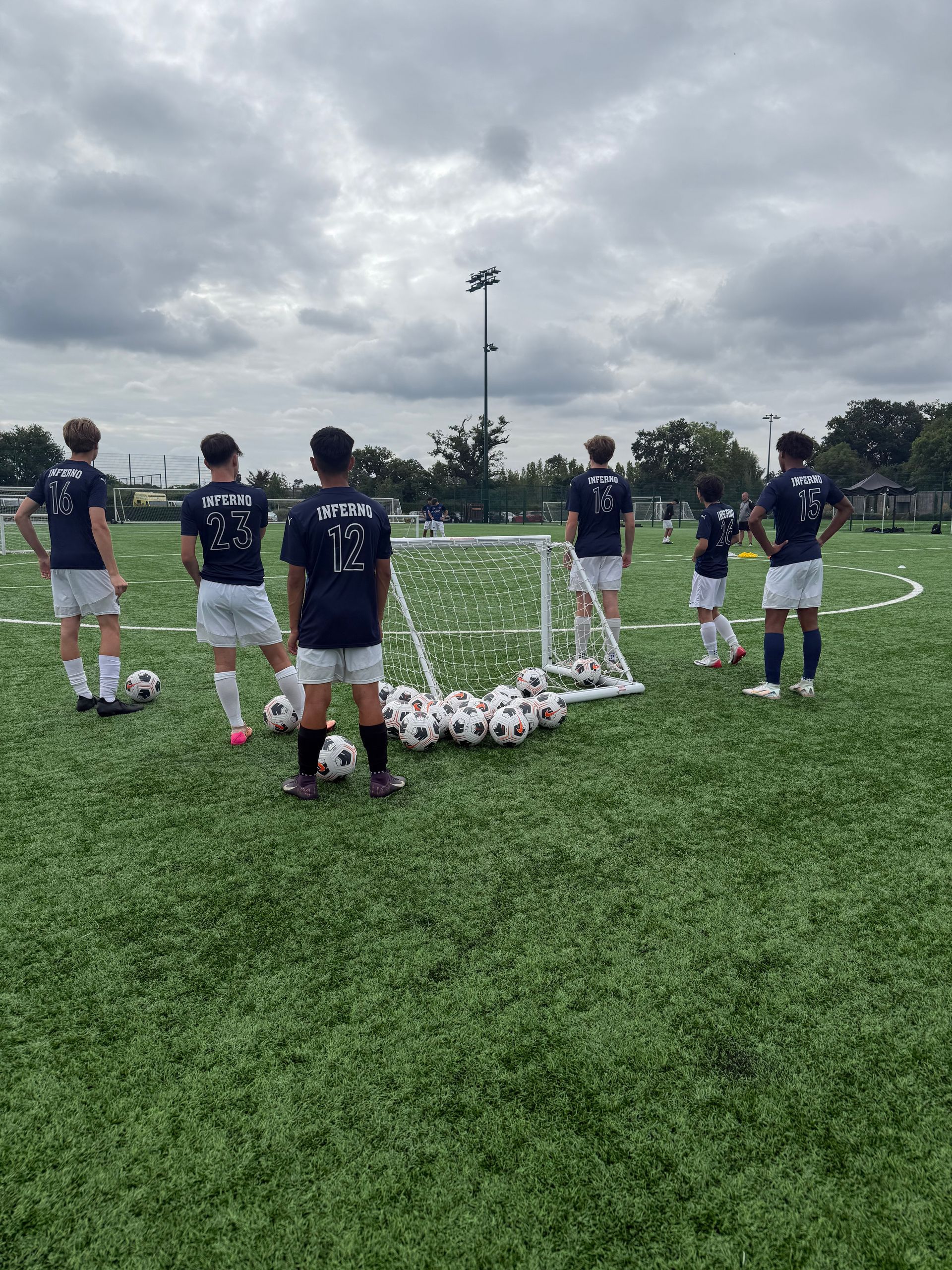 Soccer players in white shorts and dark shirts with a goal and soccer balls on a green field.