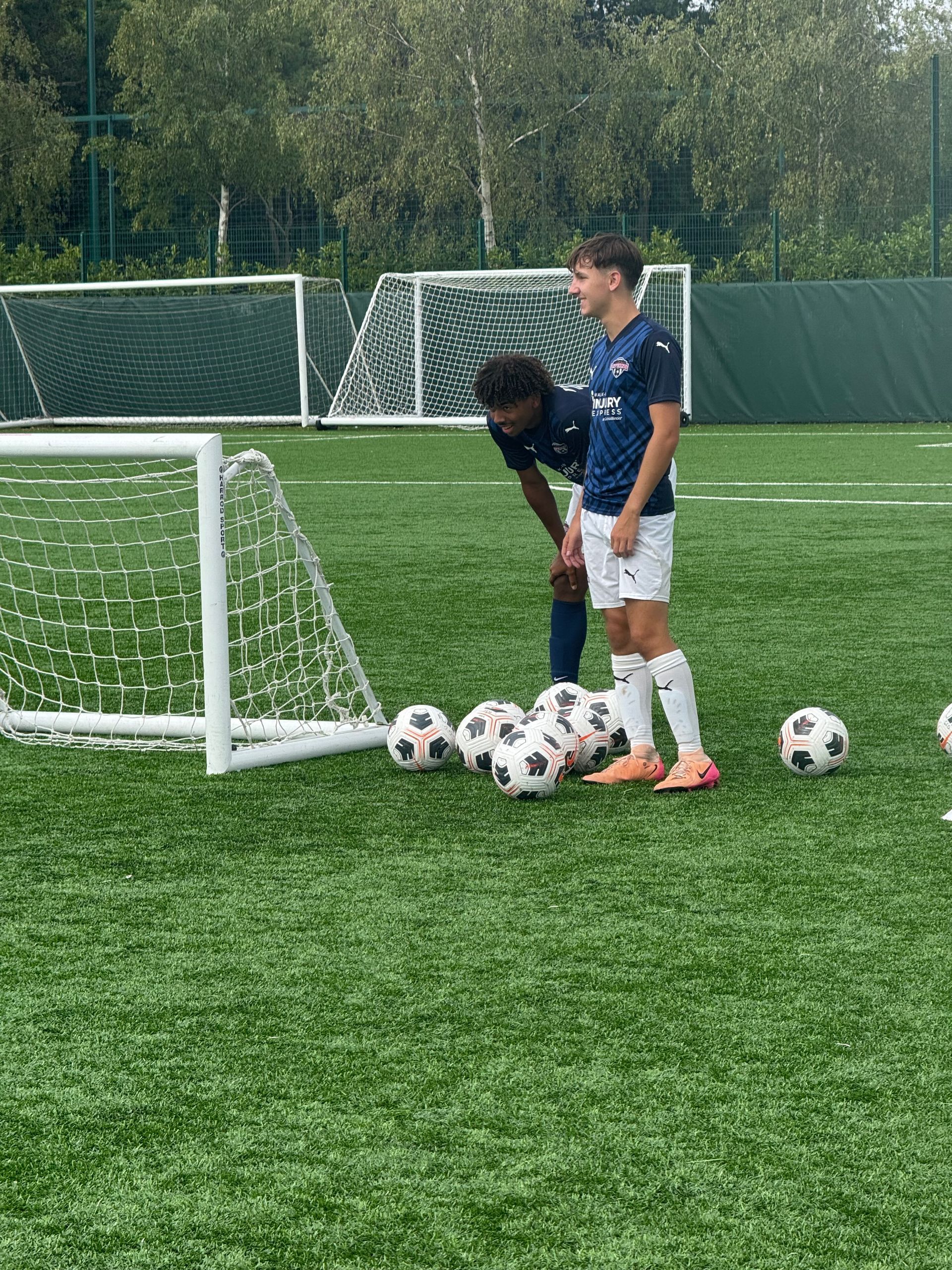 Two soccer players on a green field, one is leaning over a ball. Several soccer balls. A small white goal is to the left.