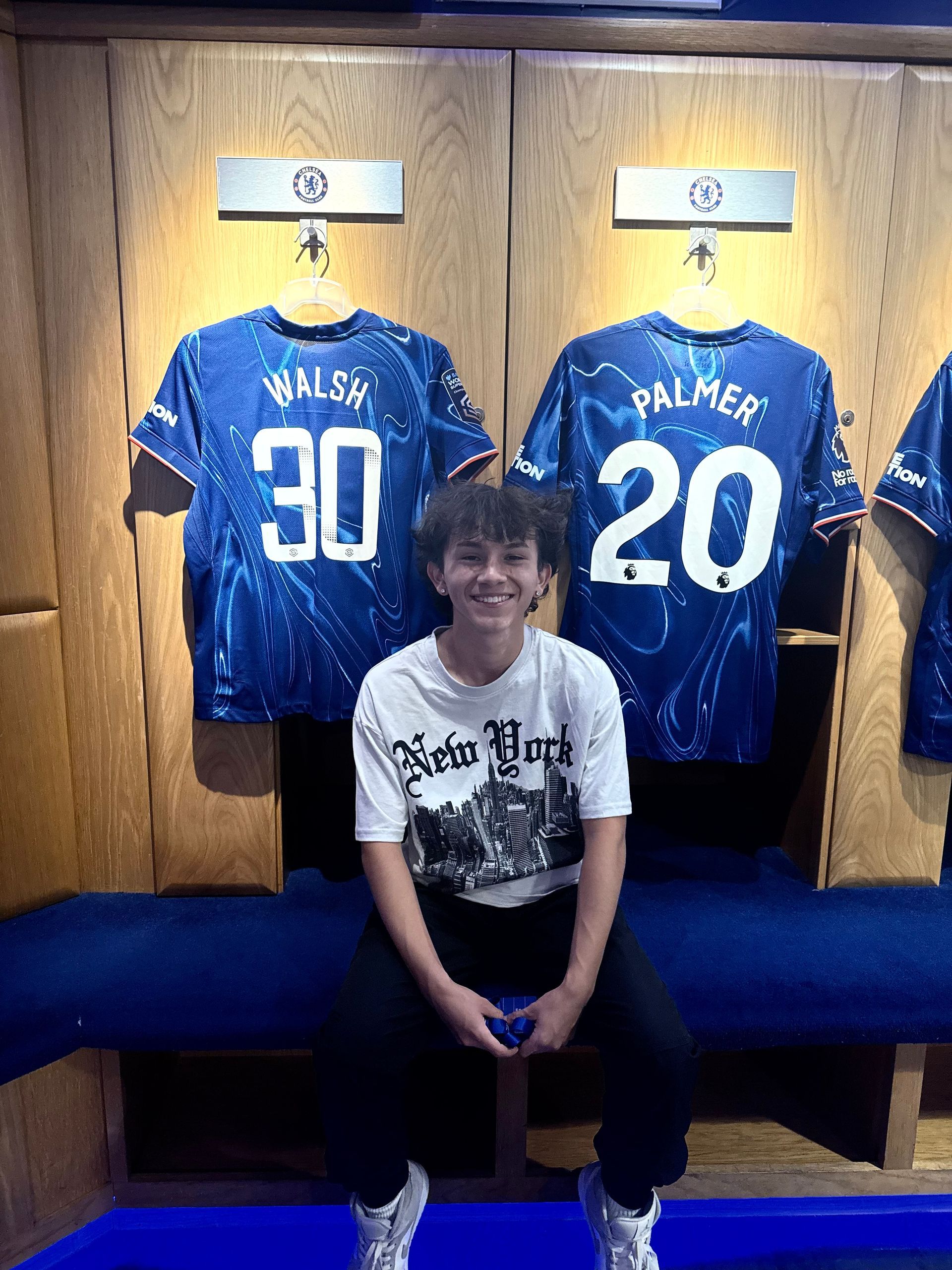 Young person smiles, sits in a Chelsea FC locker room. Blue jerseys (Walsh, 30; Palmer, 20) hang behind.