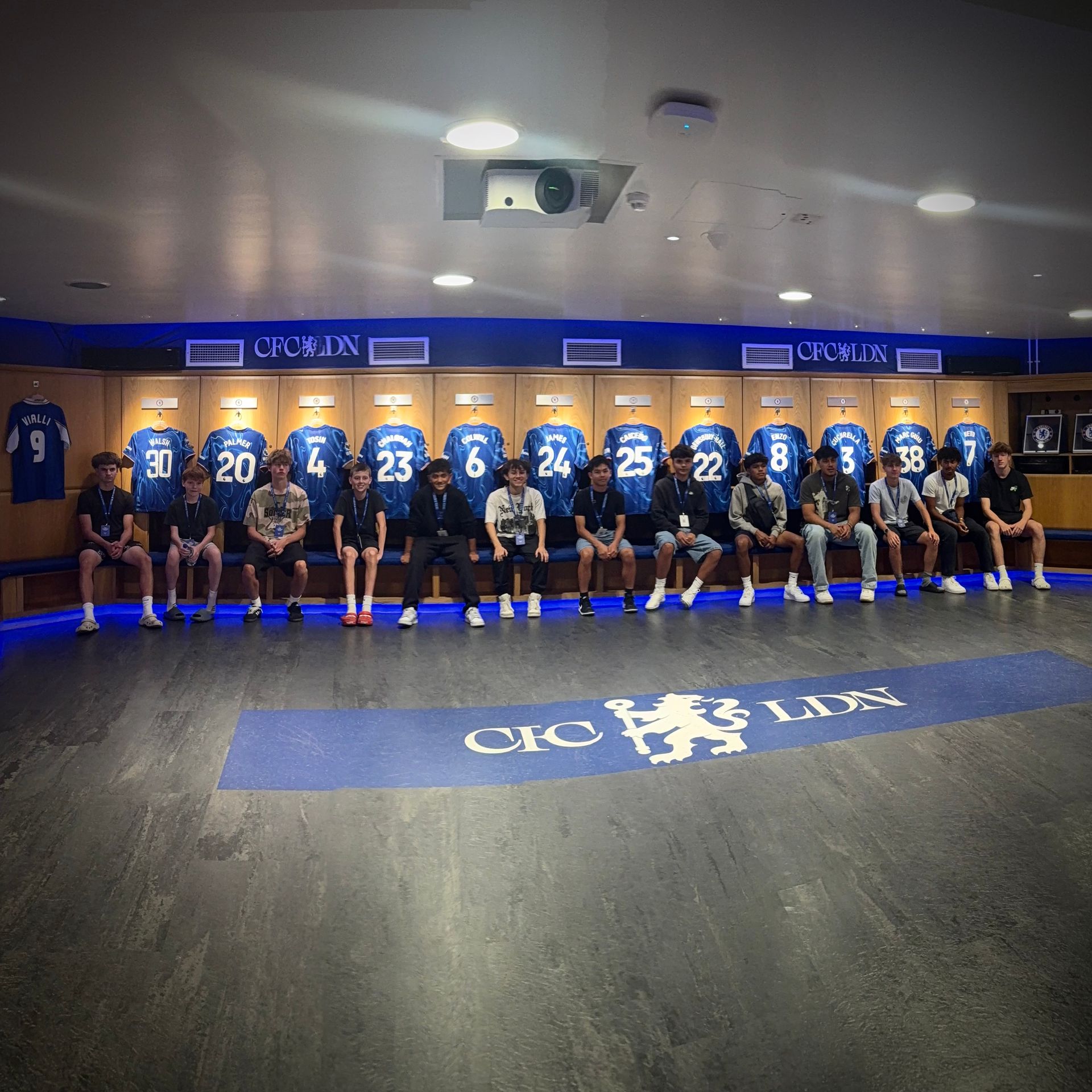 Players in blue jerseys seated in a locker room with blue accents and jerseys hanging above.
